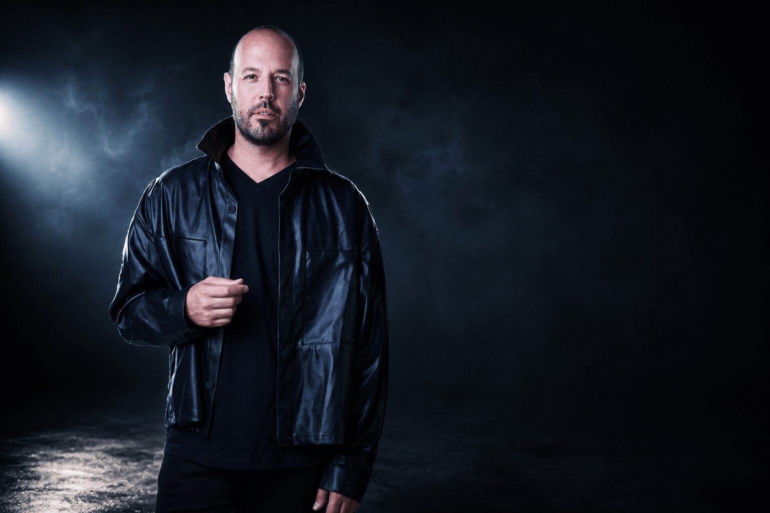 Man with a beard wearing a black leather jacket stands against a dark background.