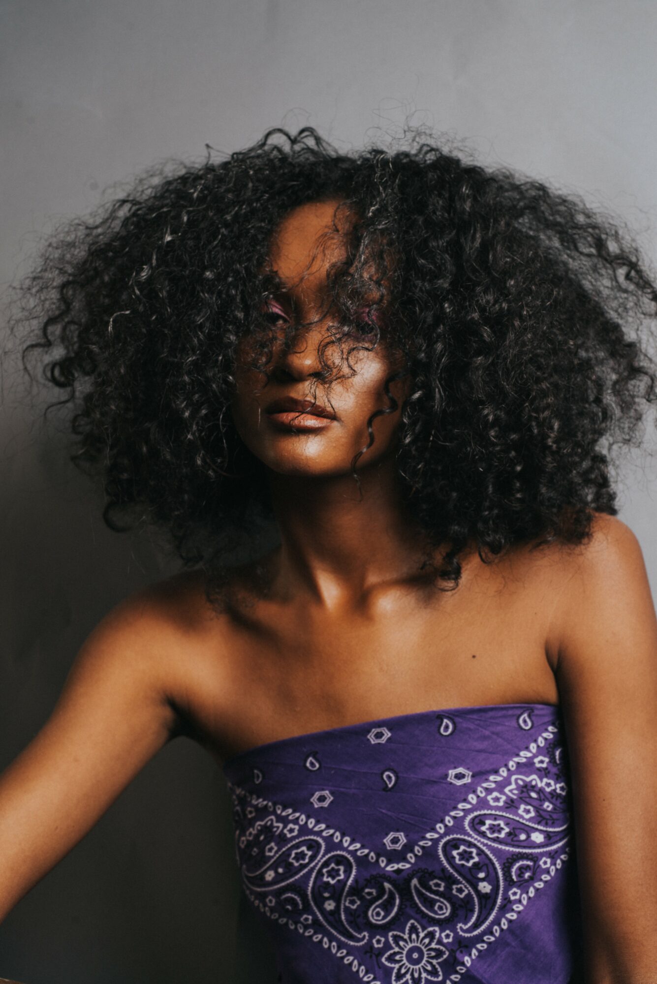 Portrait of a woman with curly hair wearing a purple strapless top, against a plain background.