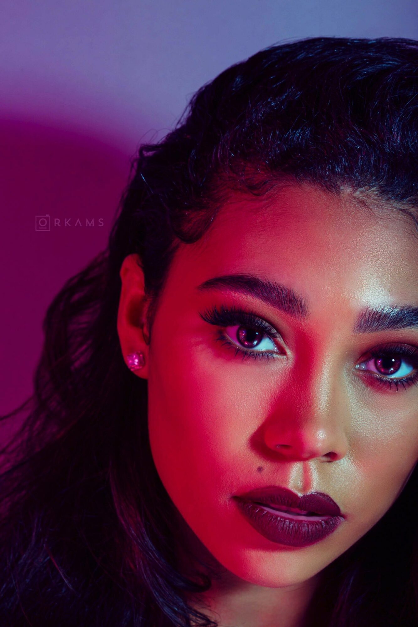 Close-up of a woman's face with makeup, dark hair, and earrings, against a purple background.