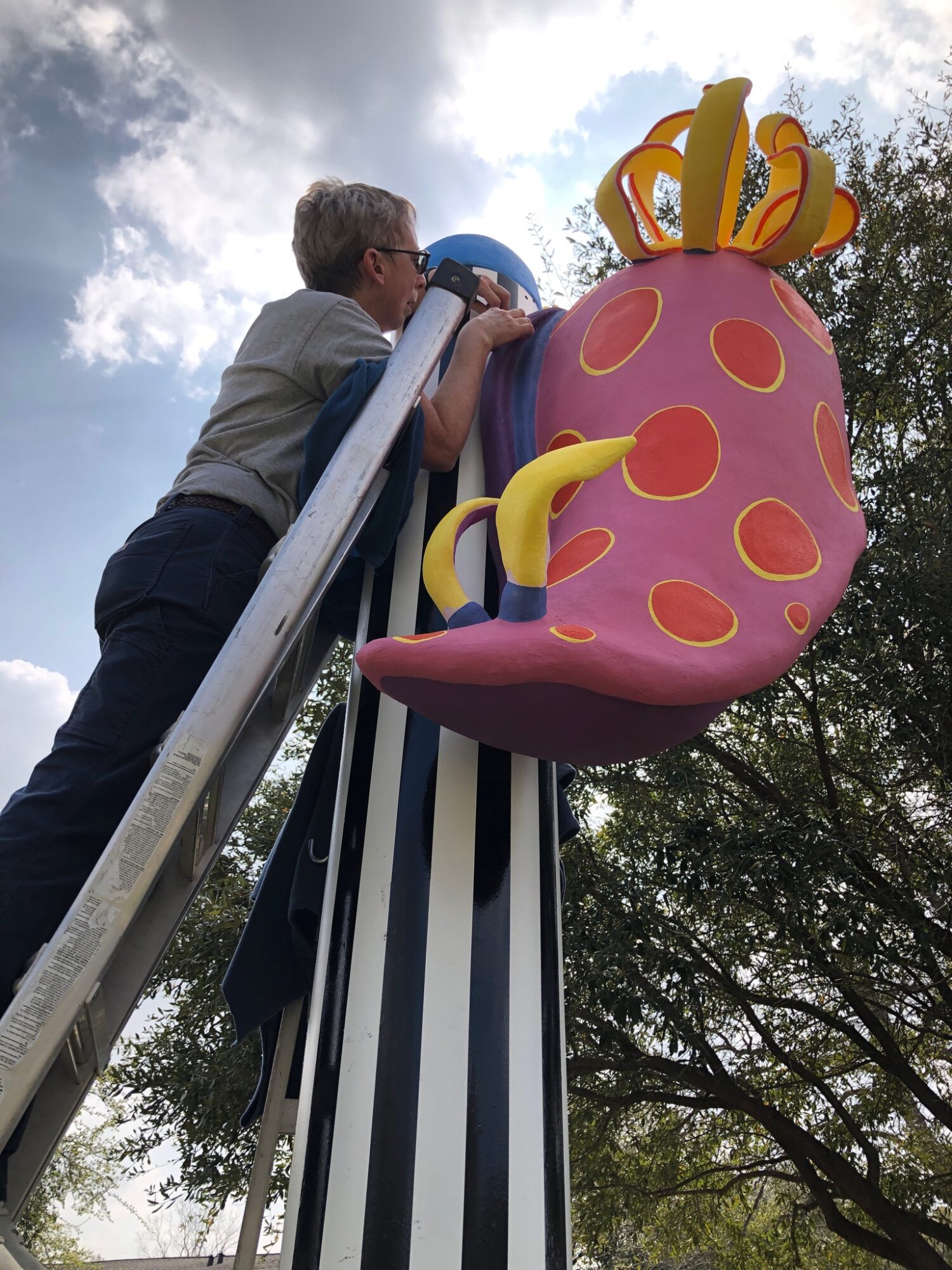 Child on a ladder reaching a large, colorful cupcake sculpture with a crown on top, outdoors with trees and cloudy sky.