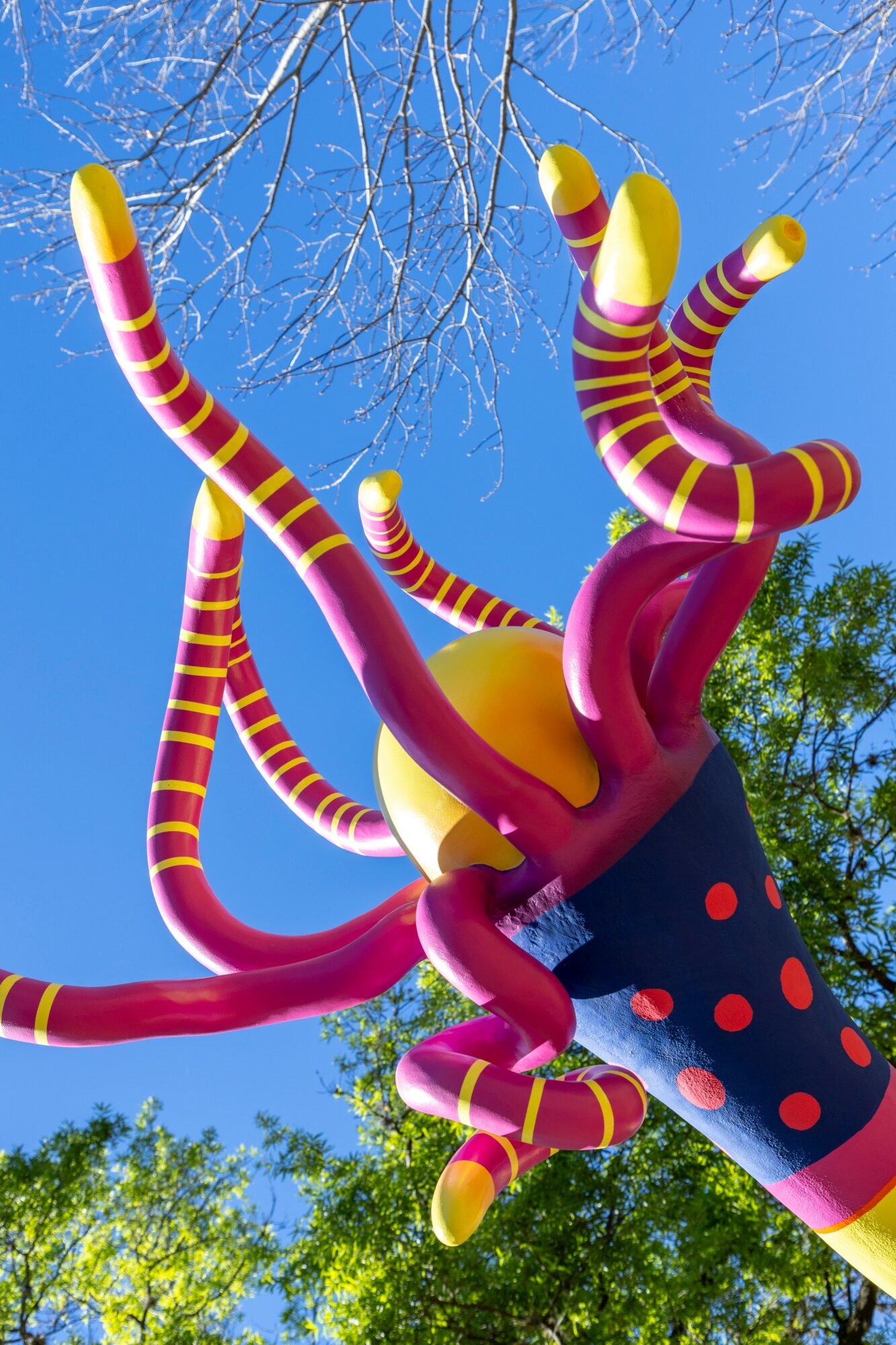 Colorful, striped, tentacle-like sculpture with a black body and red dots, set against a blue sky and green trees.