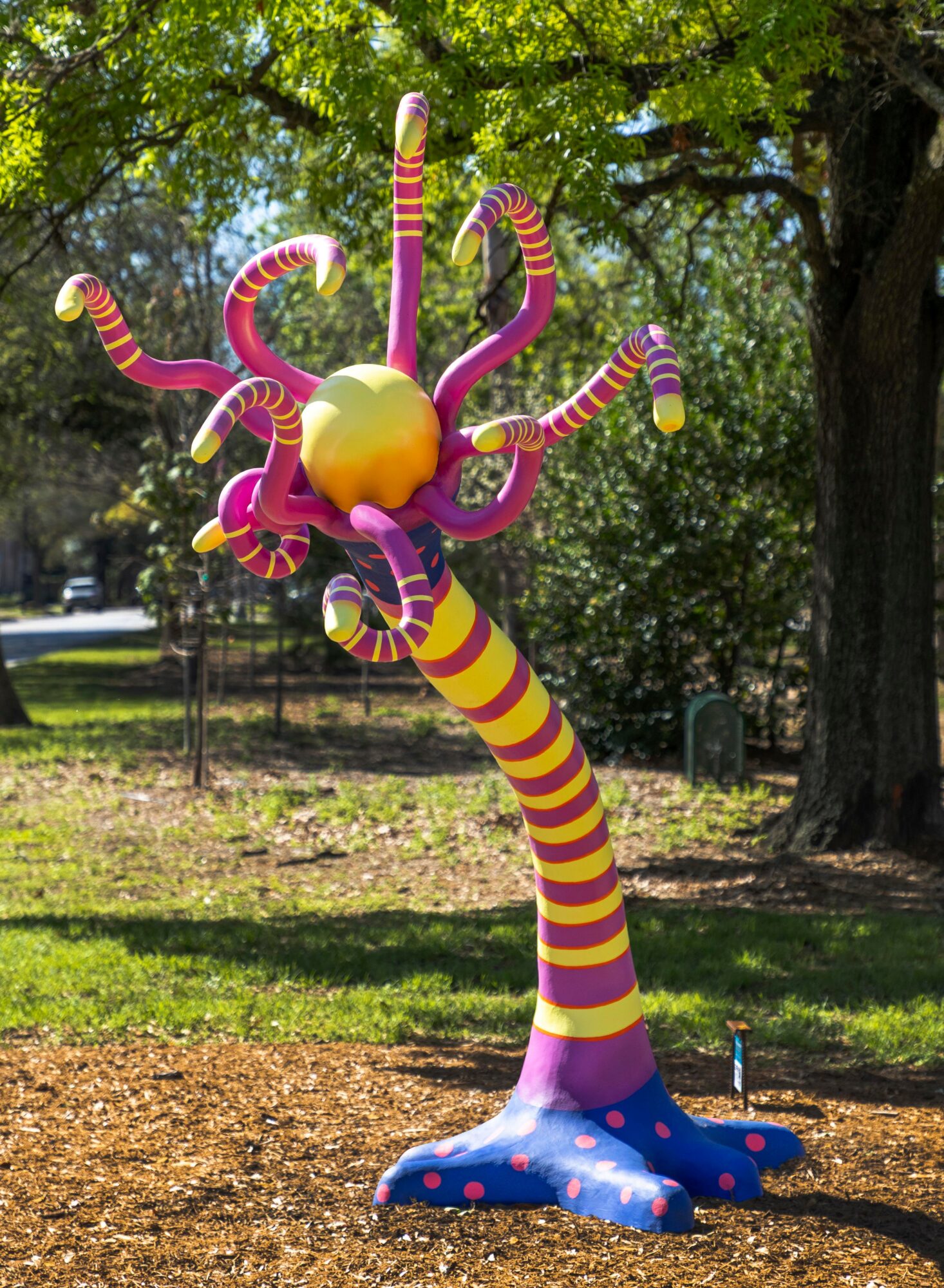Colorful abstract sculpture resembling a tree with striped, tentacle-like branches and a spherical top, in a park setting.