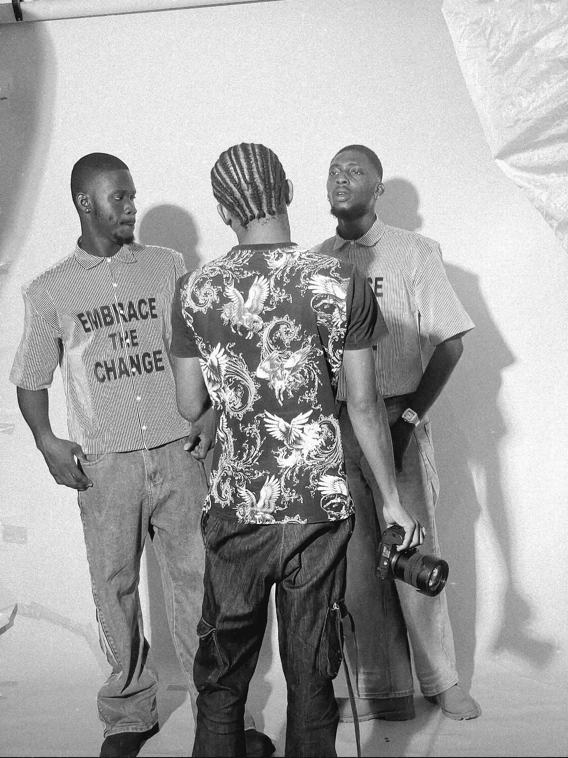 Three men standing and talking indoors, one with a camera, two facing forward, one with back to camera, plain background.