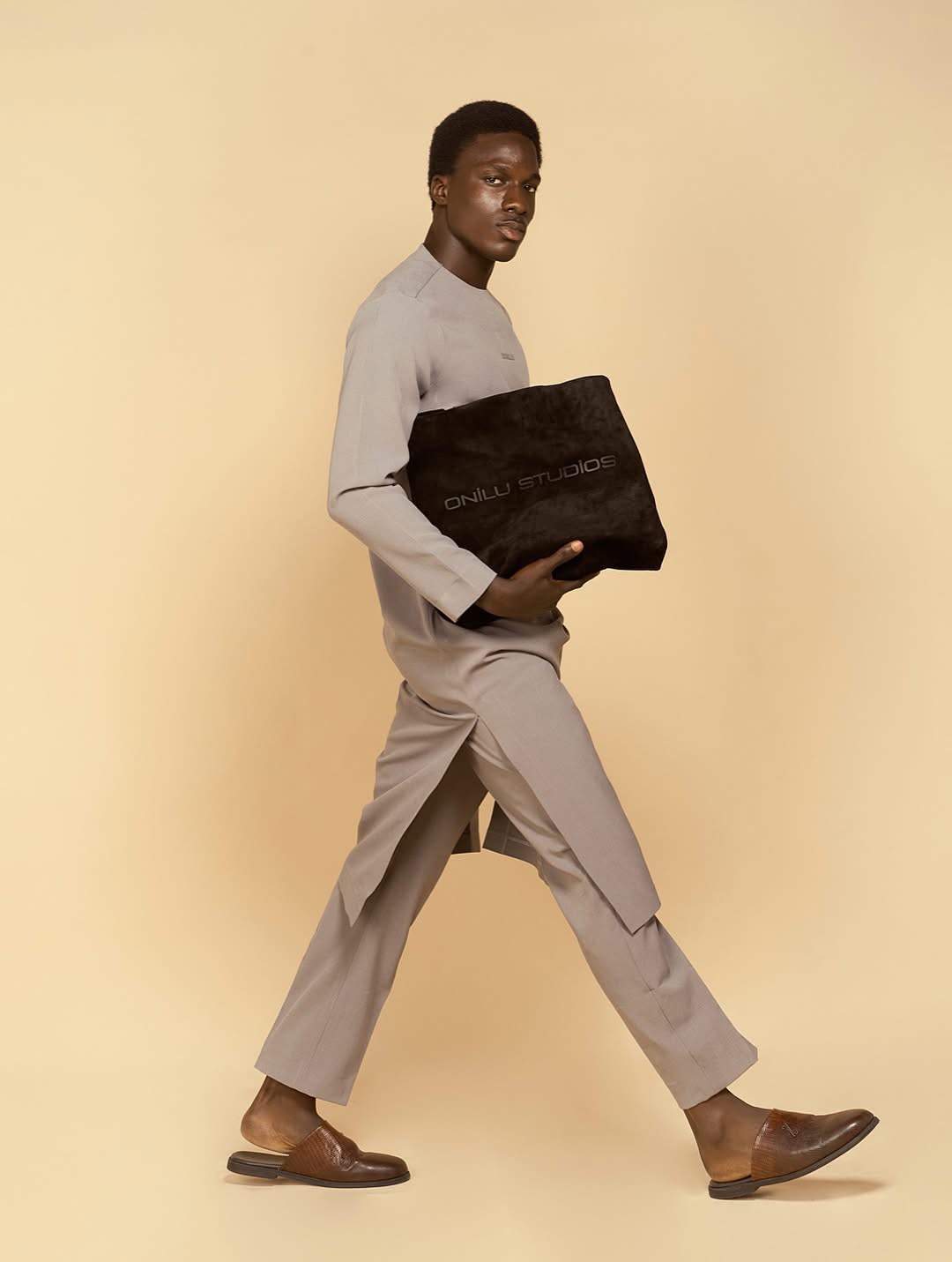Person walking with a black bag, wearing beige pants and a light gray top, against a beige background.