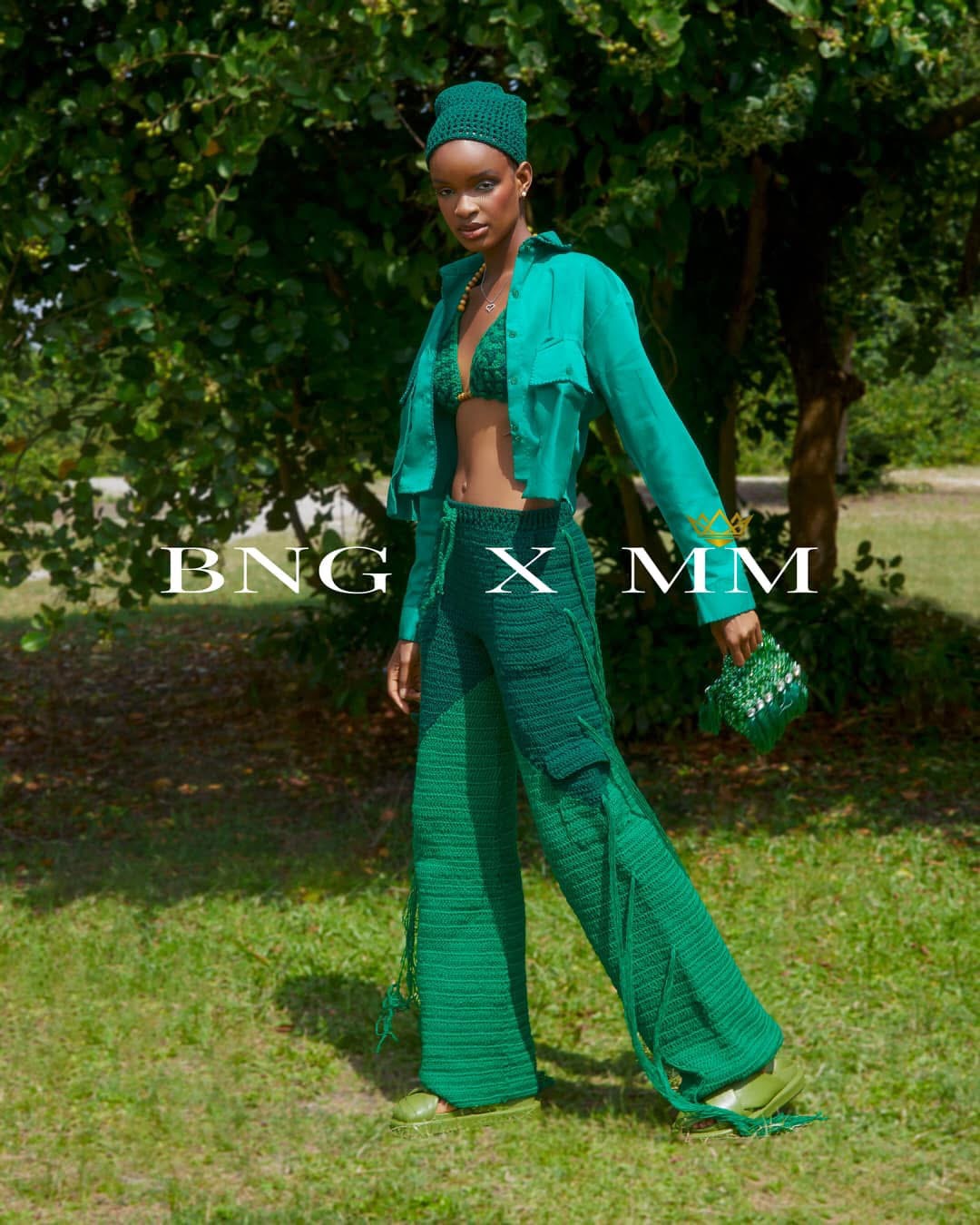 Woman standing outdoors in green outfit, holding a bag, with trees in the background.