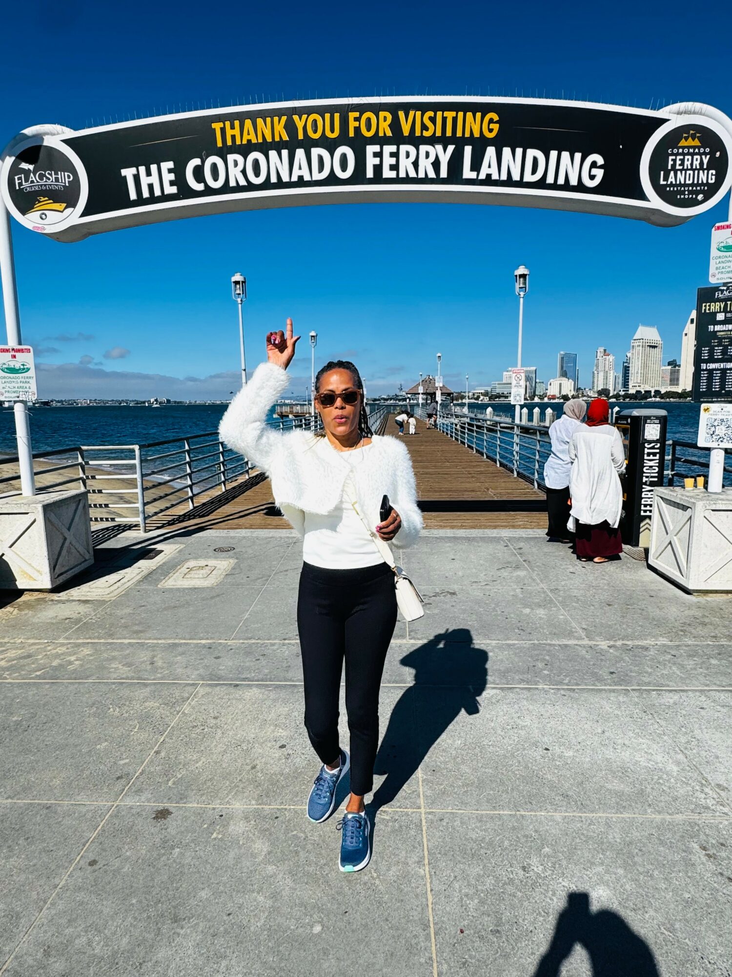 Woman standing under a sign at Coronado Ferry Landing, holding up her right hand, with water and buildings in the background.