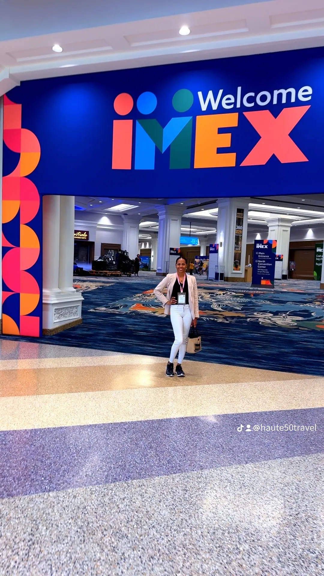 Person standing in front of a large IMEX welcome sign at a conference center, with blue carpet and white columns.