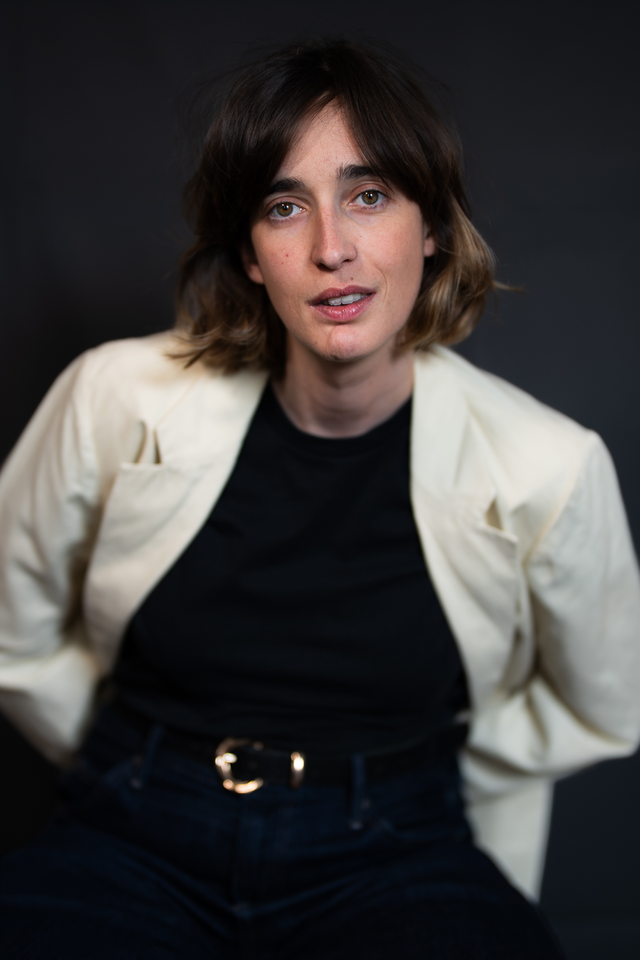 Woman with shoulder-length brown hair, wearing a white blazer and black top, against a dark background.