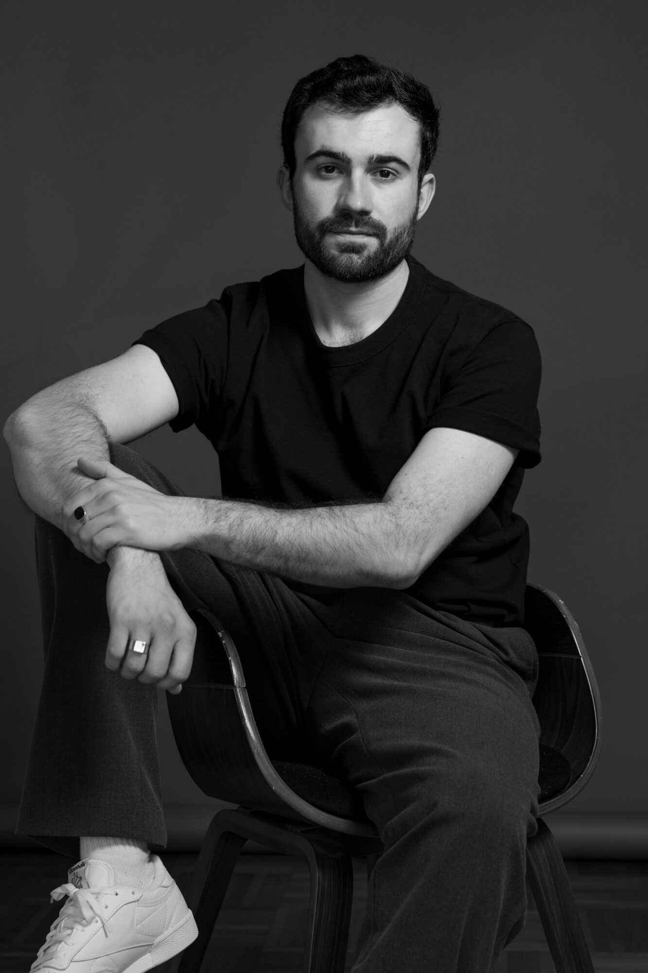 Man with beard and short hair sitting on a chair, wearing a black t-shirt and pants, against a plain background.