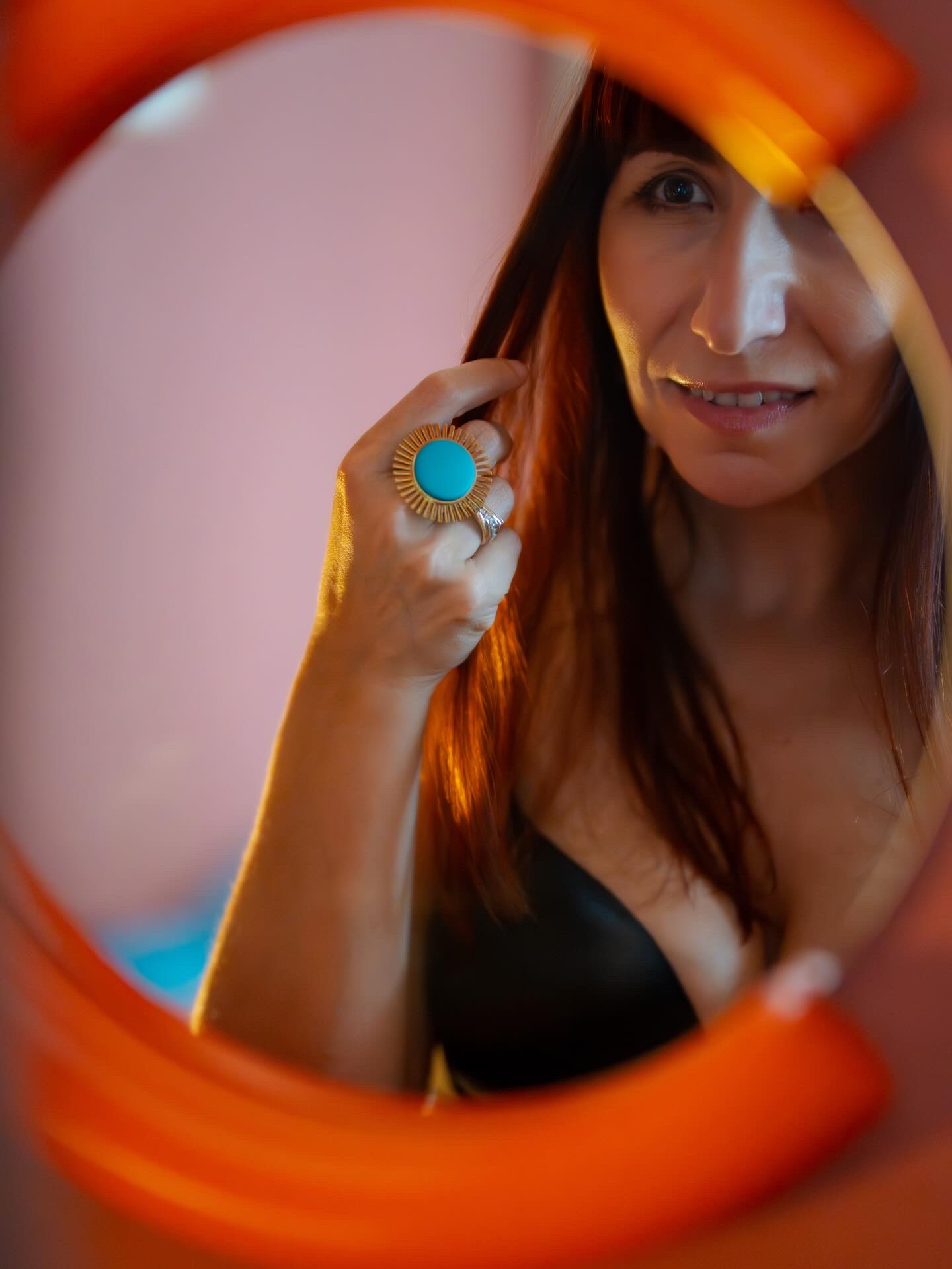Woman with long hair looking into a mirror, wearing a large ring with a turquoise stone, smiling slightly.