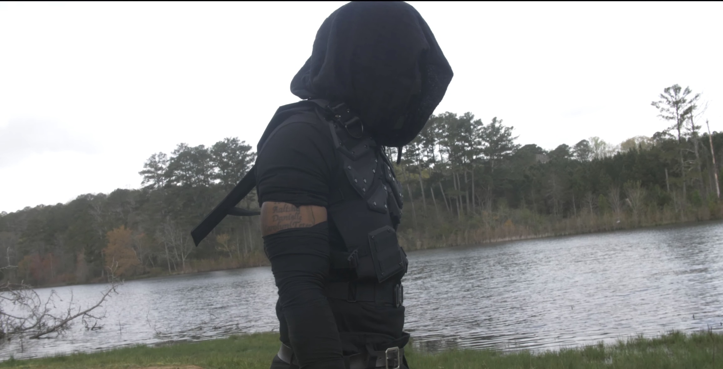 Person wearing a hooded jacket standing near a body of water with trees in the background.