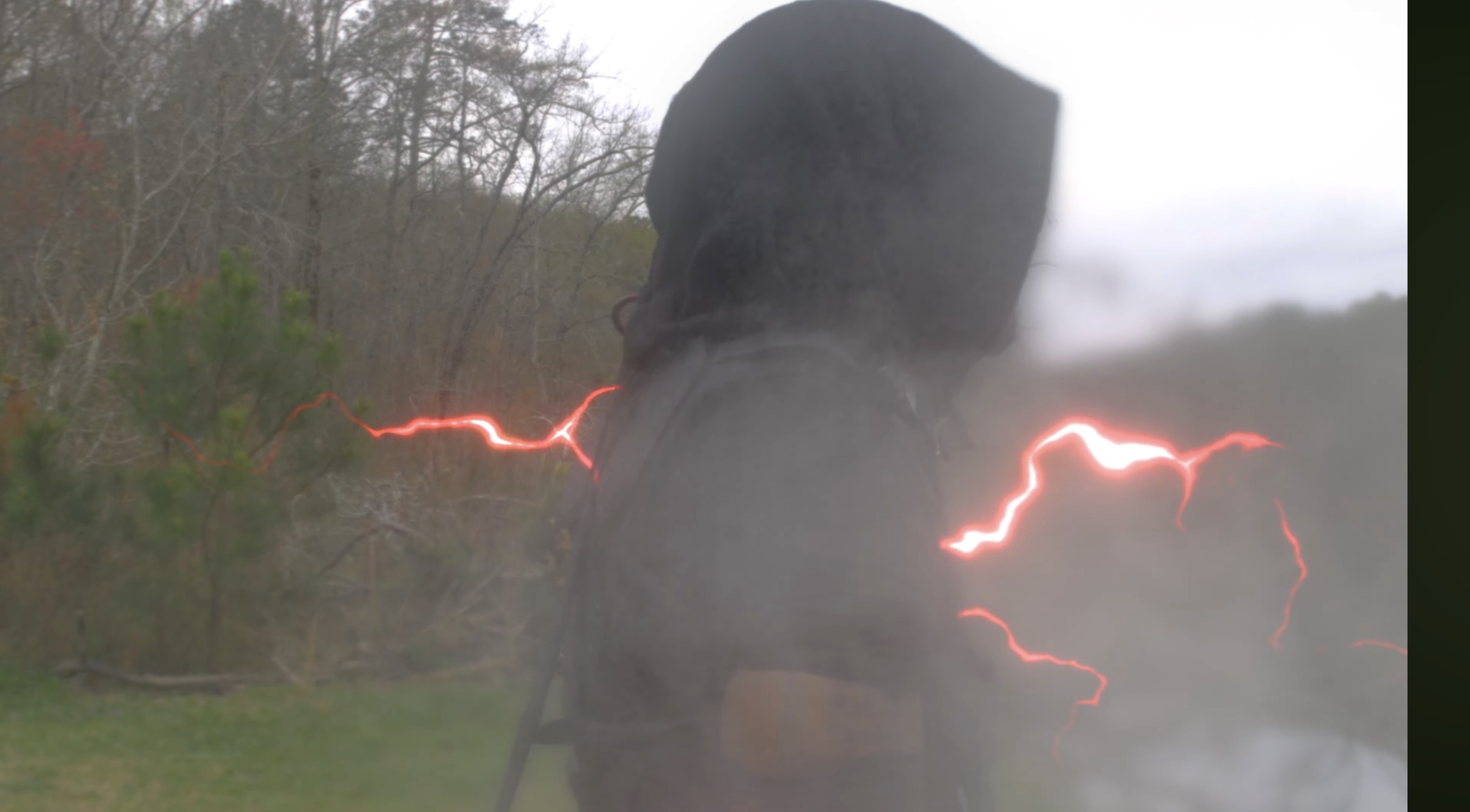 Person with dark hooded clothing outdoors, lightning in the background, cloudy sky, trees, and grass, foggy atmosphere.