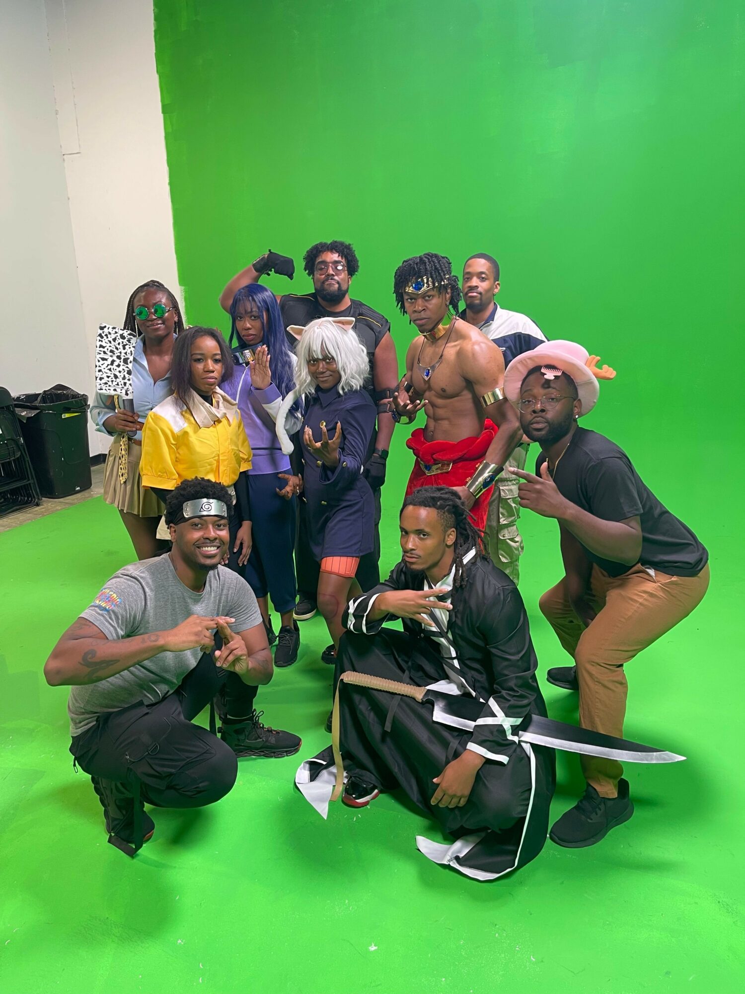 Group of people in front of a green screen, some sitting and some standing, with one person in a wheelchair, all smiling.