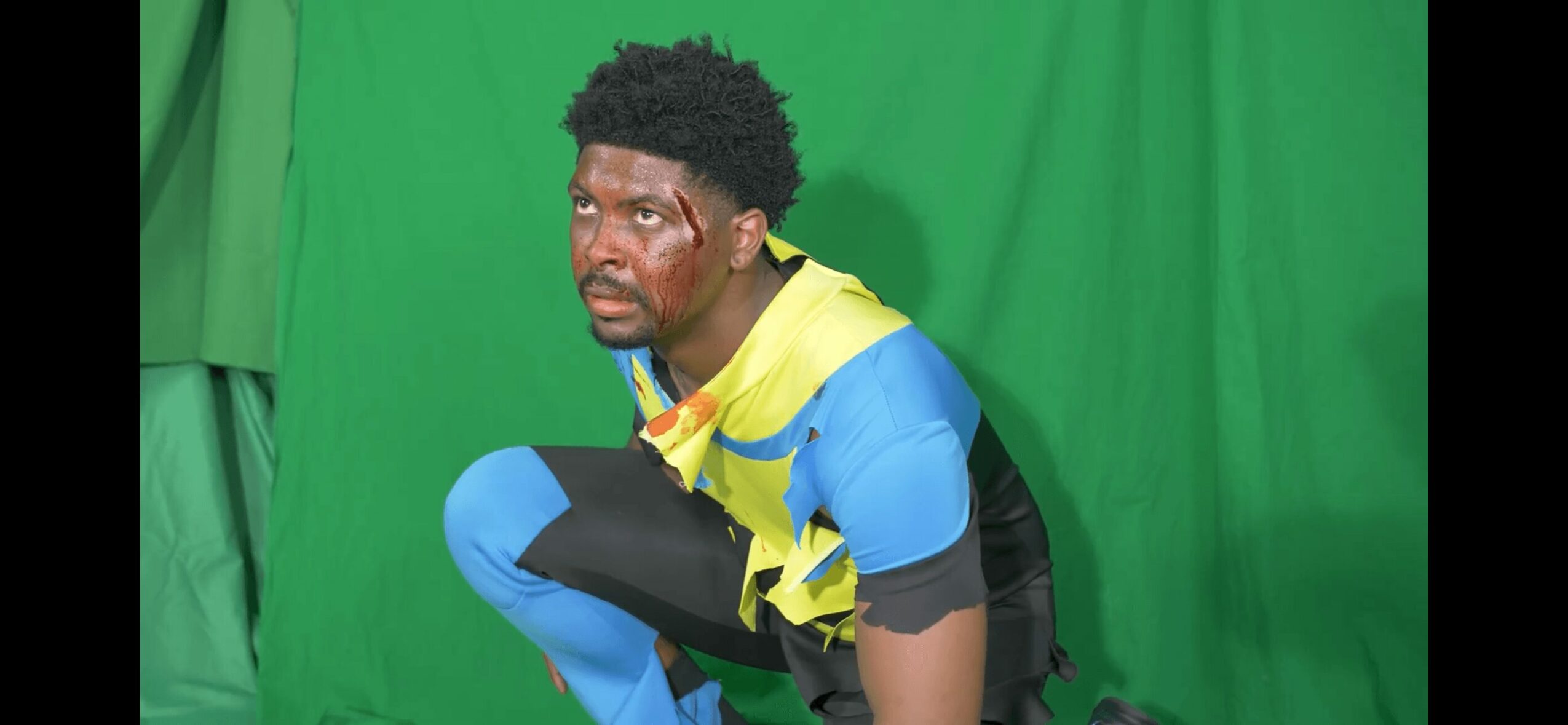 Person crouching in front of a green screen, wearing a yellow and blue sports outfit, with a serious expression.