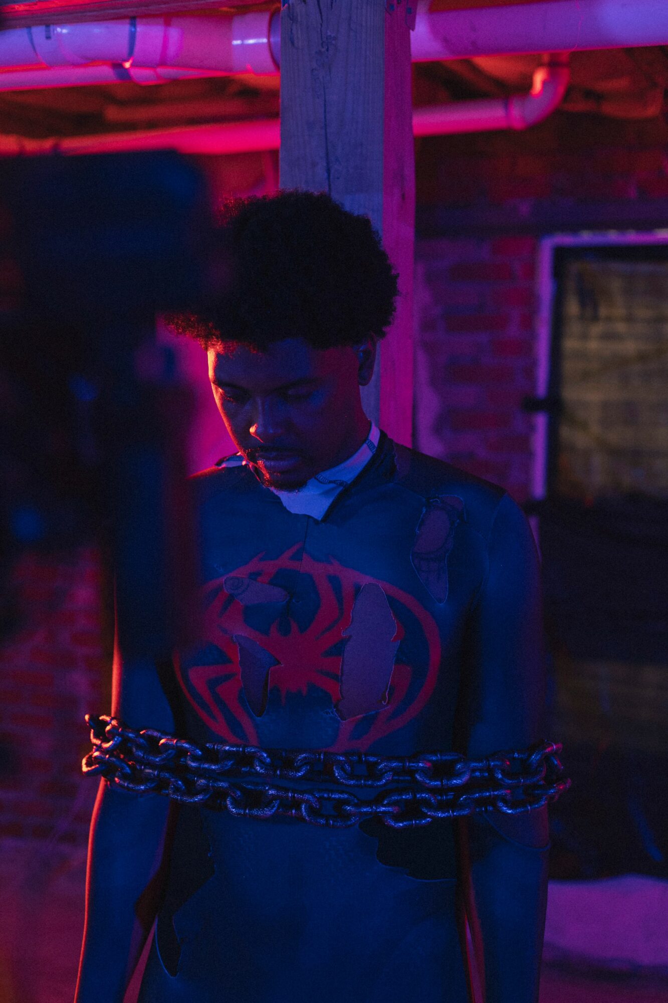 Person with curly hair in a dark room with pink and purple lighting, chained at the waist, wearing a Spider-Man shirt.