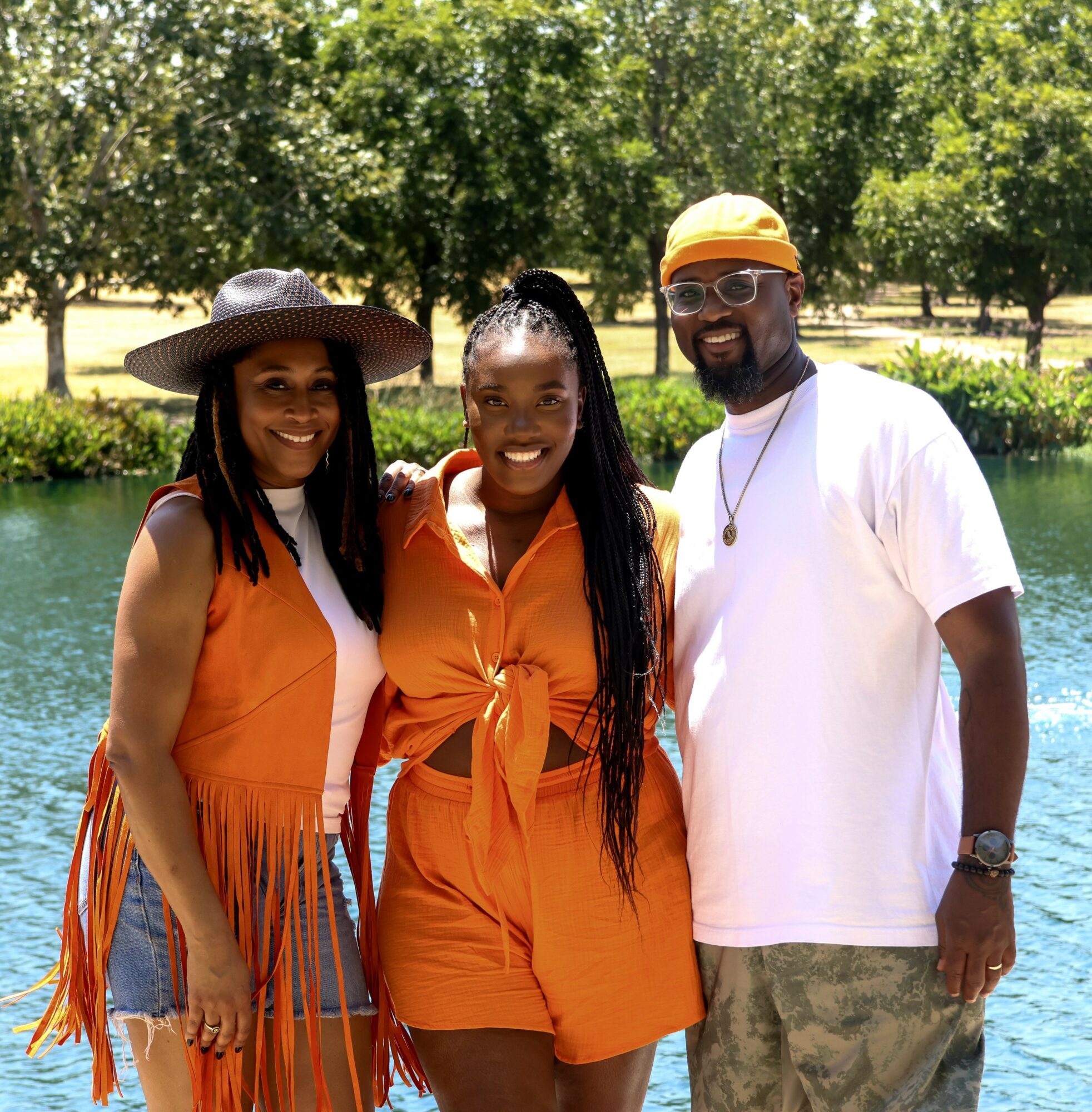 Three people standing outdoors near water, smiling, with trees in the background. Two women and one man, all dressed casually.
