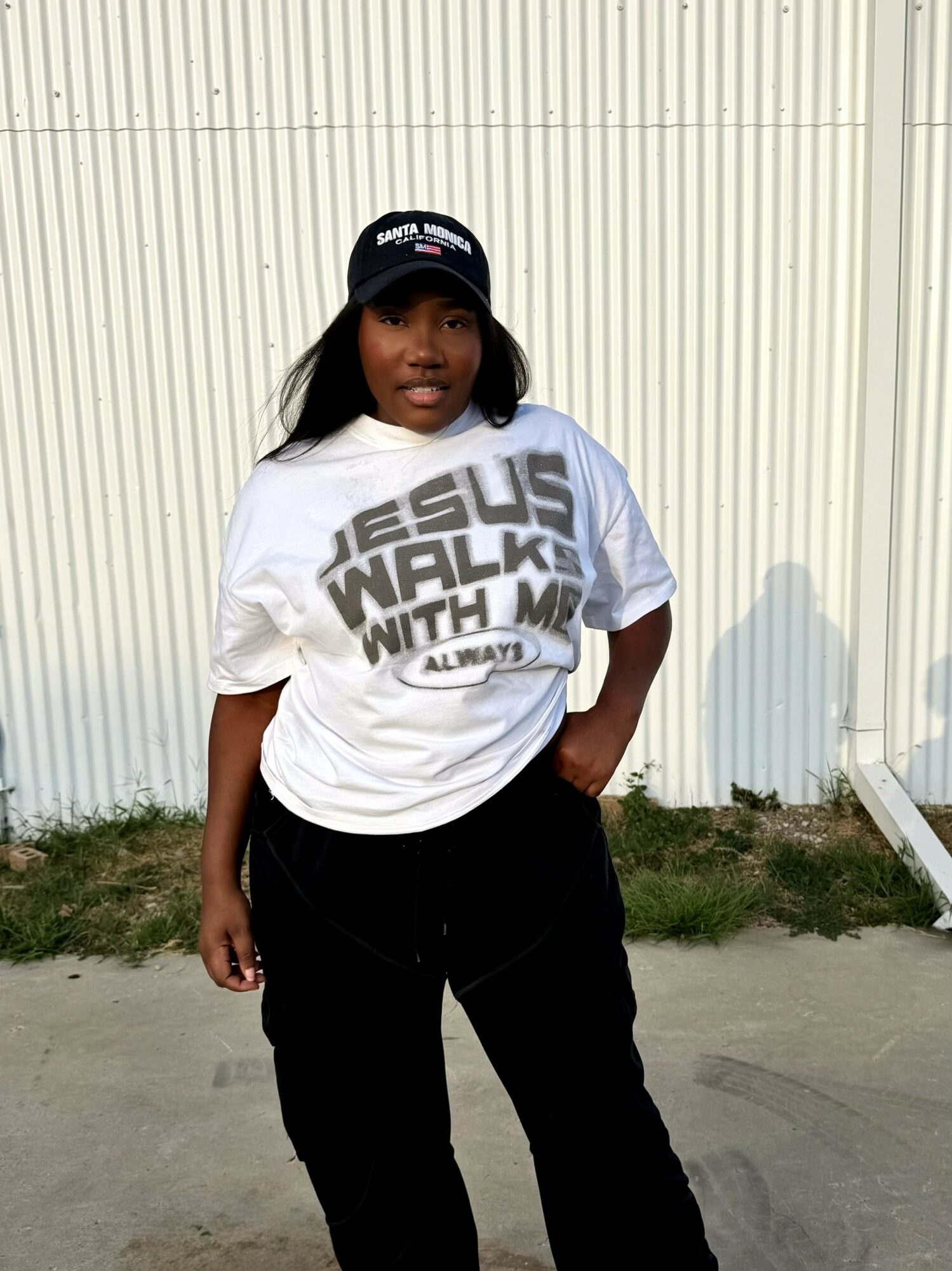 Woman standing outdoors in front of a white wall, wearing a white T-shirt with text, black pants, and a cap.