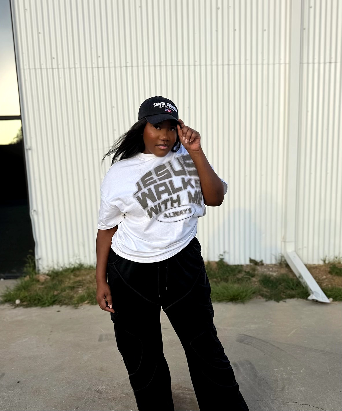 Woman standing outdoors in front of a white wall, wearing a white t-shirt, black pants, and a black cap, adjusting her cap.
