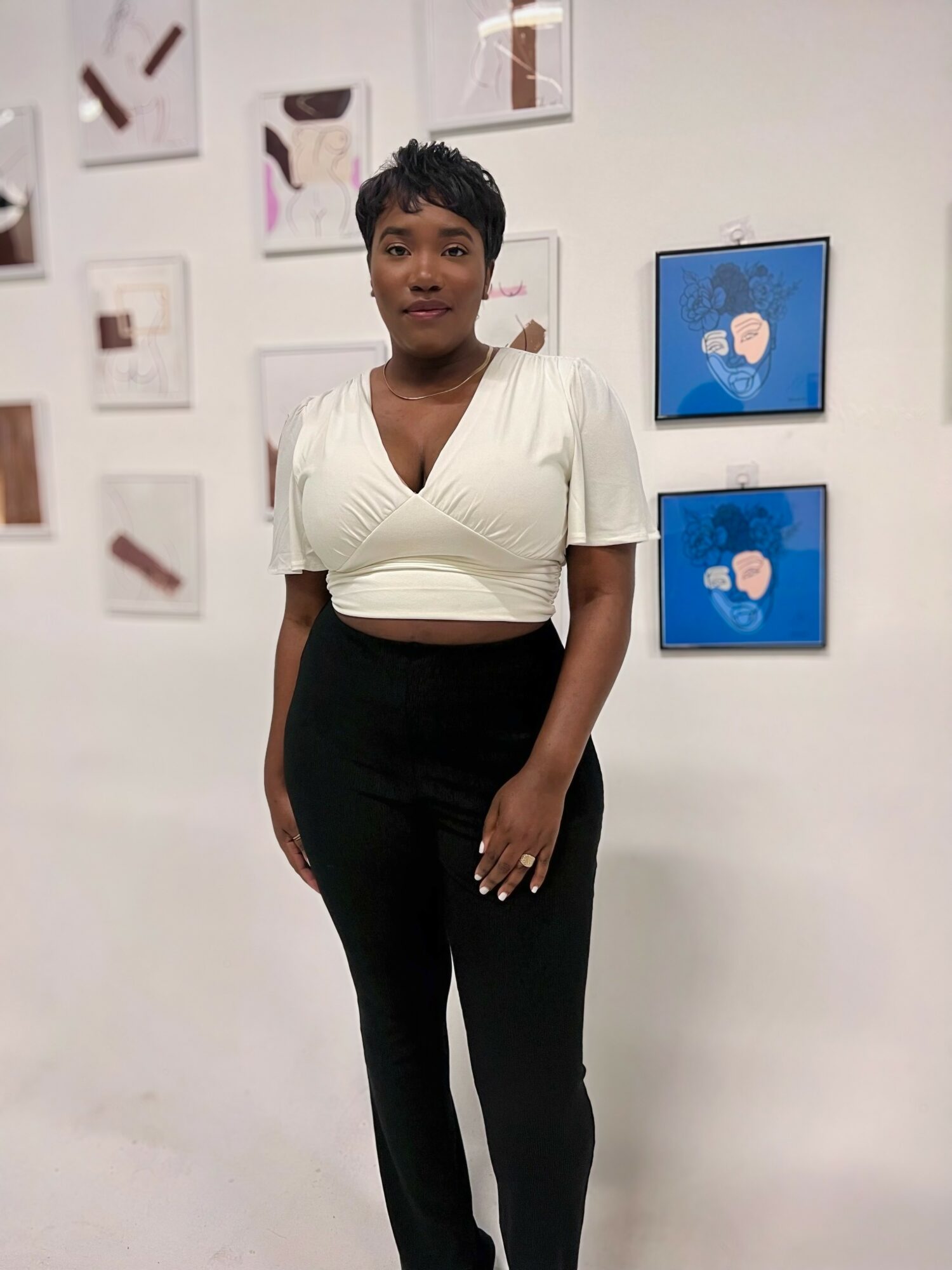 Woman standing in front of wall with framed artwork, wearing white top and black pants, looking at camera.