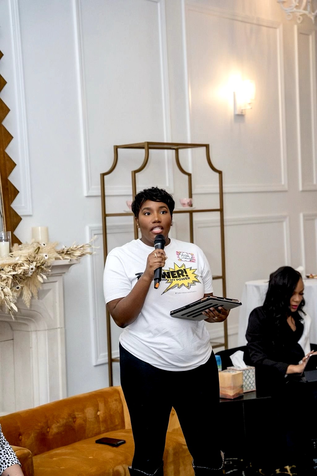 Woman holding a microphone and a clipboard, speaking at an indoor event with elegant decor.