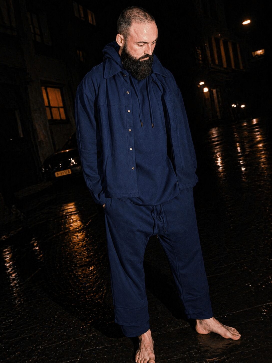 Man with a beard stands barefoot on a wet street at night, wearing a dark blue jacket and pants, looking down.