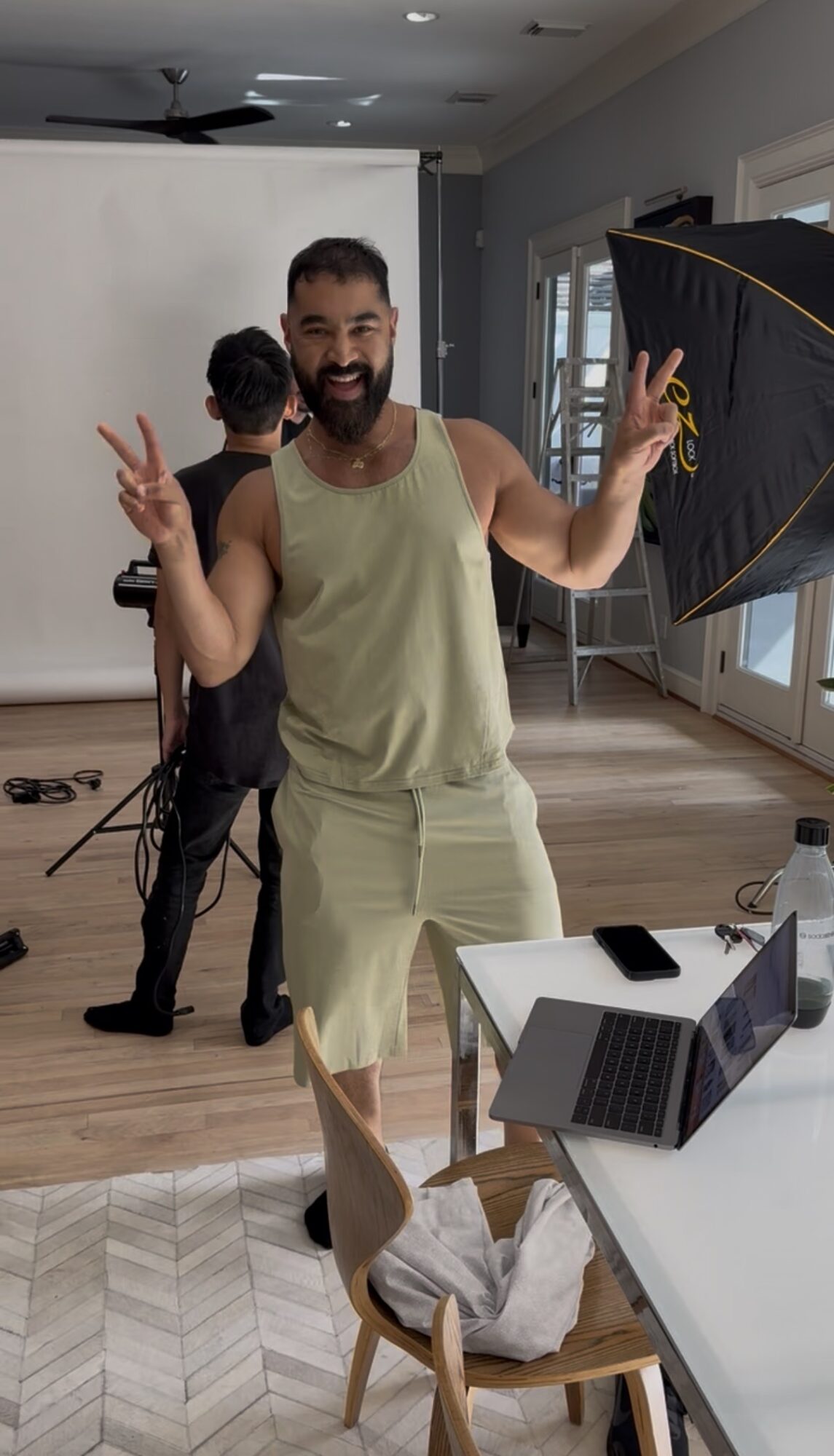 Man smiling and making peace signs indoors, with a person in black behind him, a table with a laptop, and a swan sculpture.