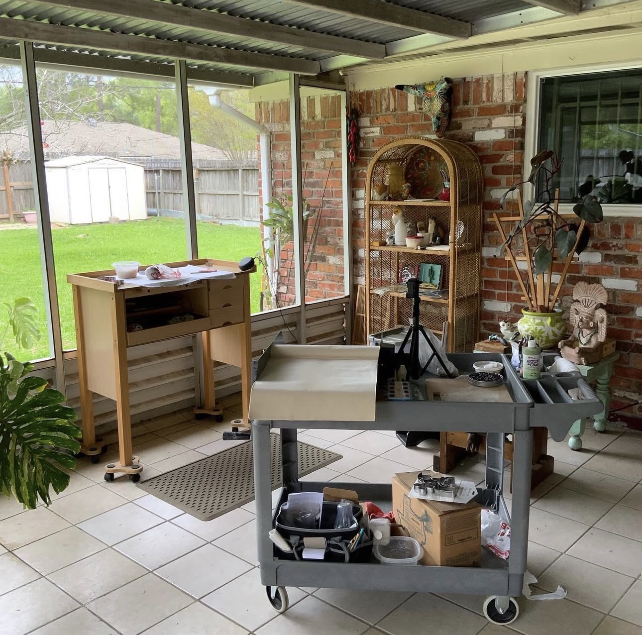 Indoor space with a cart, a wooden table, a brick wall, and a window showing a backyard with grass and trees.