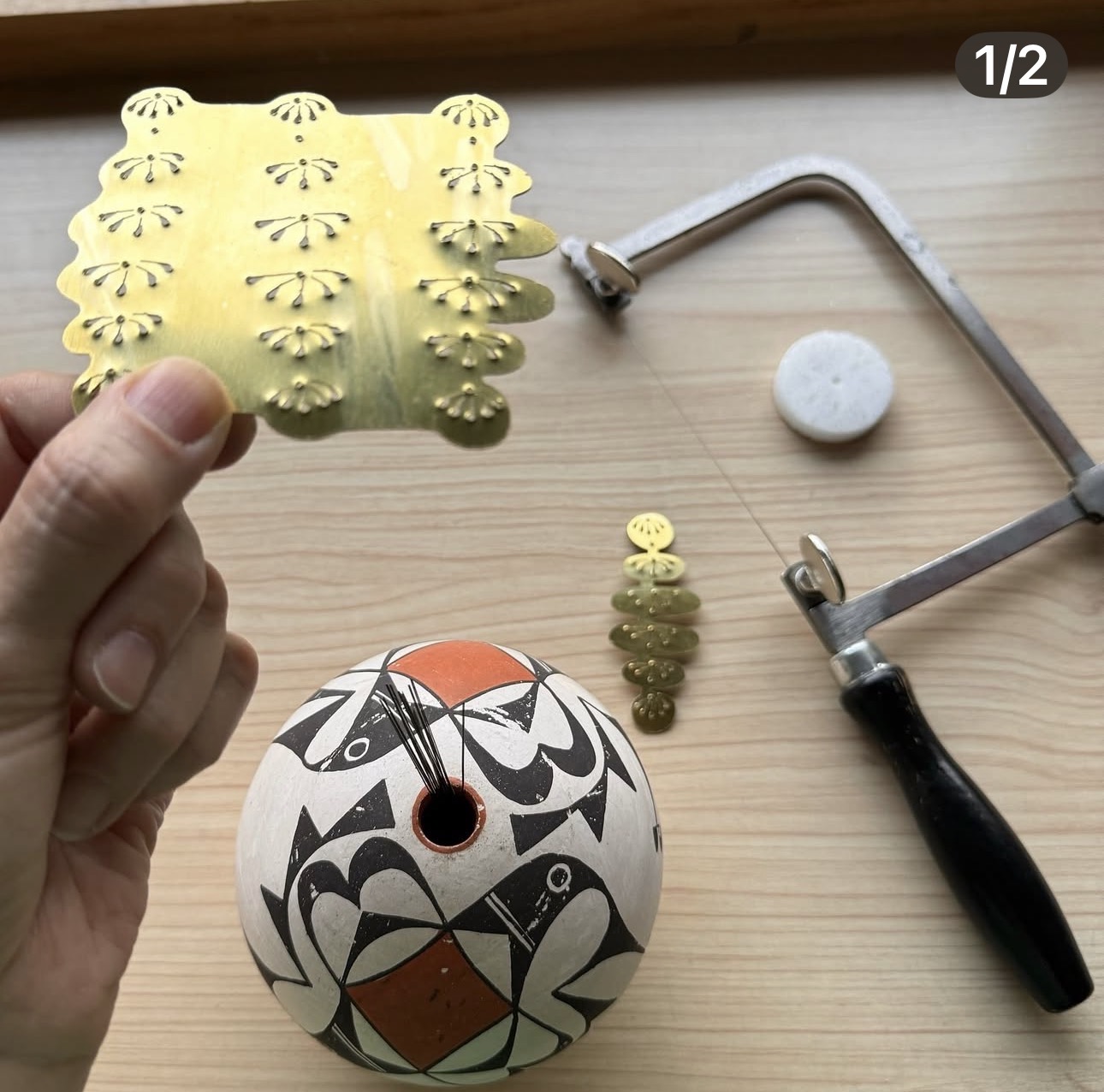 Hand holding a yellow decorative sheet with cut-out patterns, a clock with a black and white design, and small objects on a wooden surface.