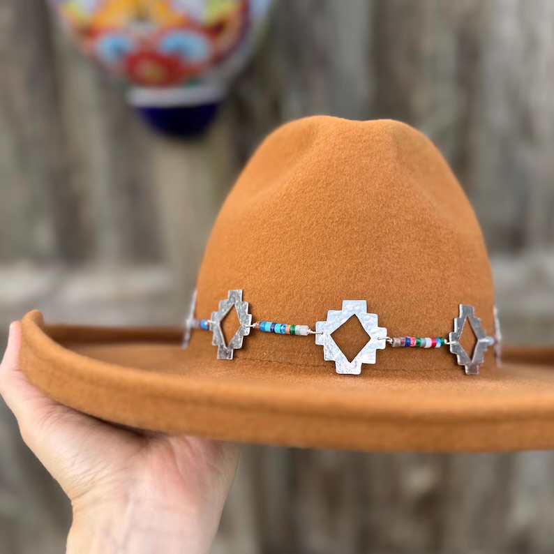 Hand holding a brown hat with a decorative band, outdoors with a wooden background.