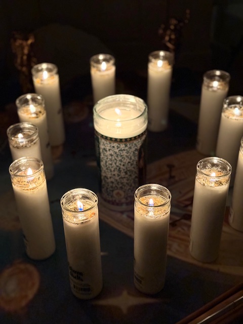 Multiple lit candles arranged in a circle on a dark surface, with a decorative glass in the center.