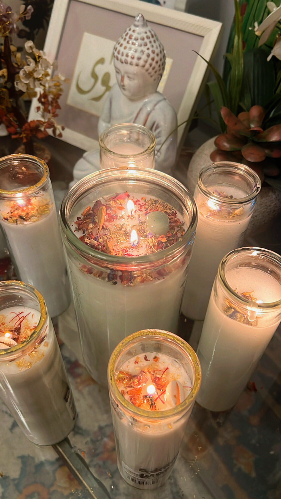Multiple lit candles and a Buddha statue in a decorative setting with plants and framed artwork behind.