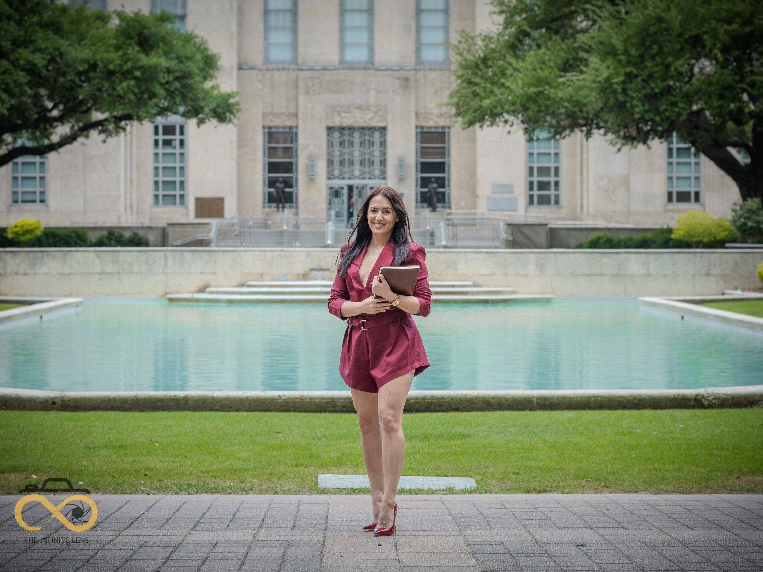 Young woman holding a book and a folder, standing in front of a fountain on a university campus.