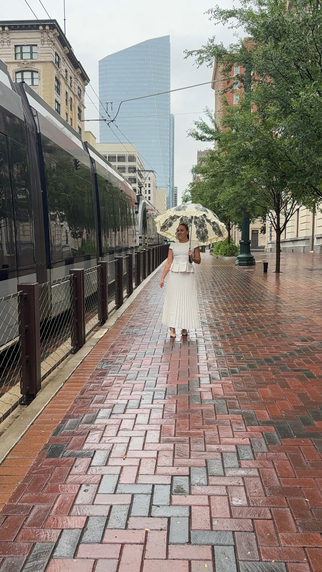 Person in white dress holding an umbrella on a brick sidewalk with buildings and trees, cityscape in background.