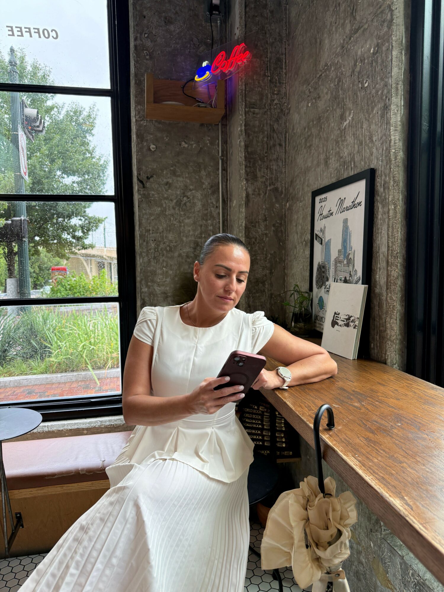 Woman in white dress sitting at a wooden table, holding a phone, with a window showing trees outside.