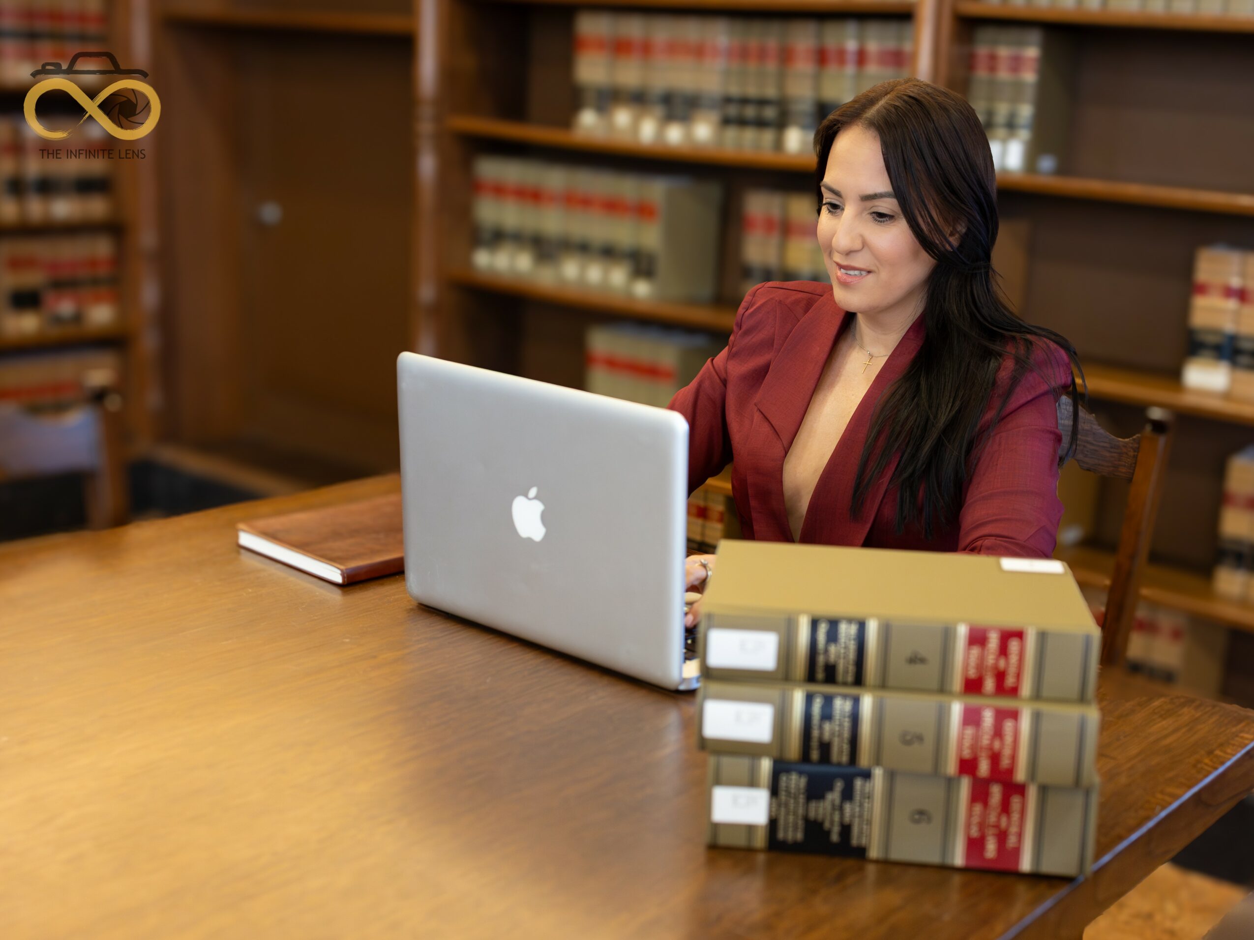 Woman in red blazer working on a laptop at a wooden table in a library, with bookshelves in background.
