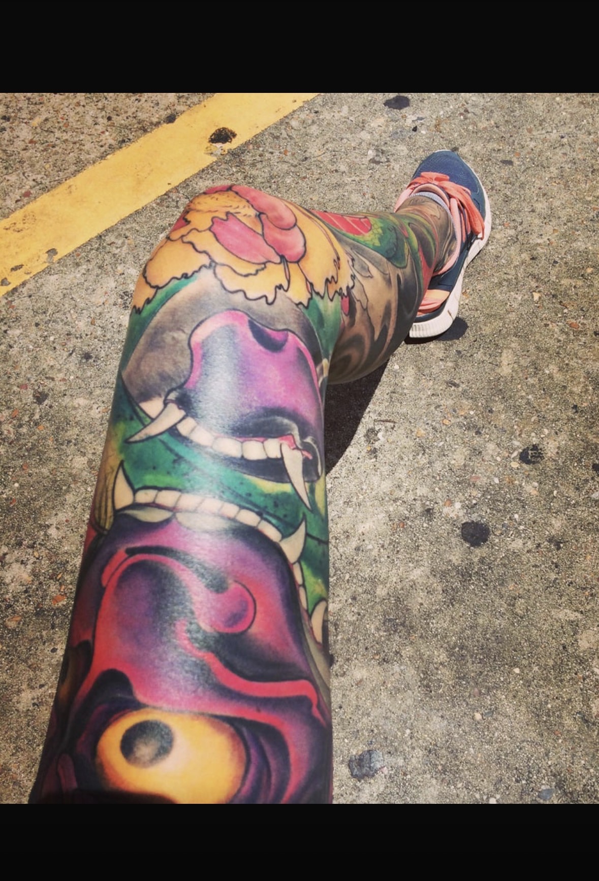 Person's leg with colorful tattoos, wearing a sneaker, on a concrete surface with a yellow line.