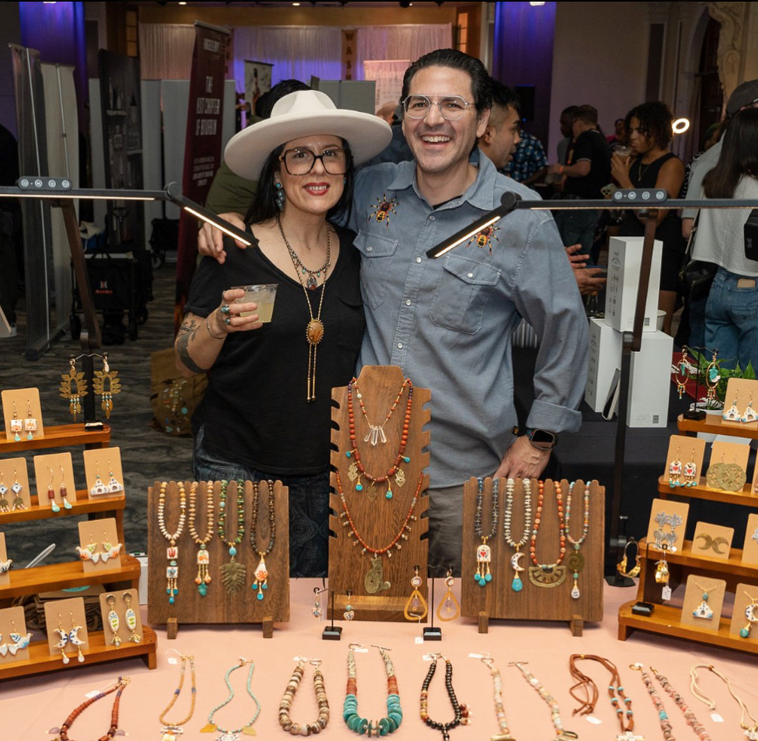 Two people smiling at a jewelry booth, one wearing a large hat, jewelry, and glasses, the other in a gray shirt, in an indoor market.