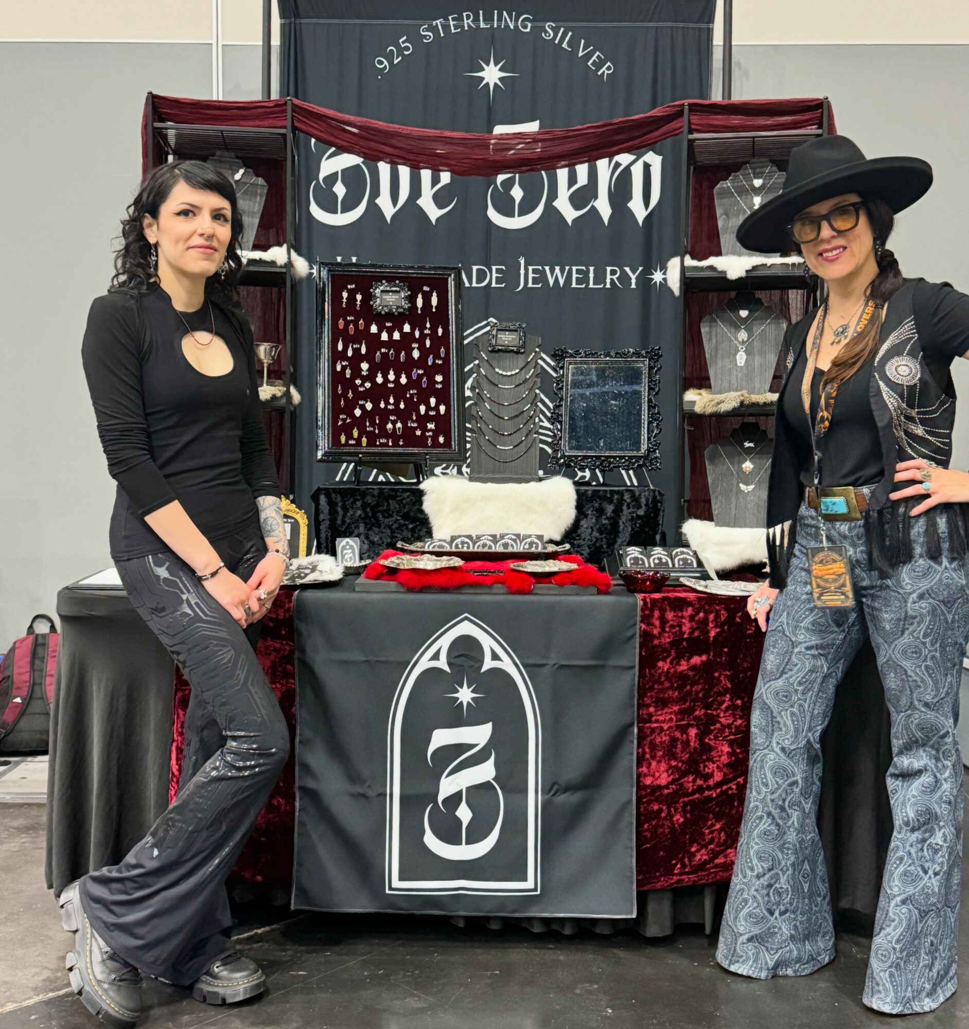 Two women stand beside a jewelry display booth with jewelry and a banner reading