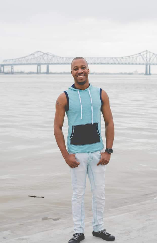 Smiling man standing on beach with bridge in background, wearing sleeveless hoodie, light pants, and sneakers.