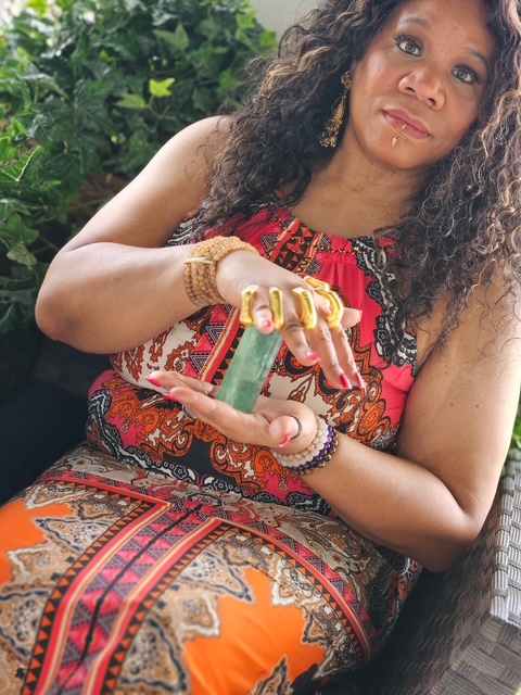 Woman with curly hair wearing colorful patterned dress holding a glass with a drink, outdoors with greenery in background.