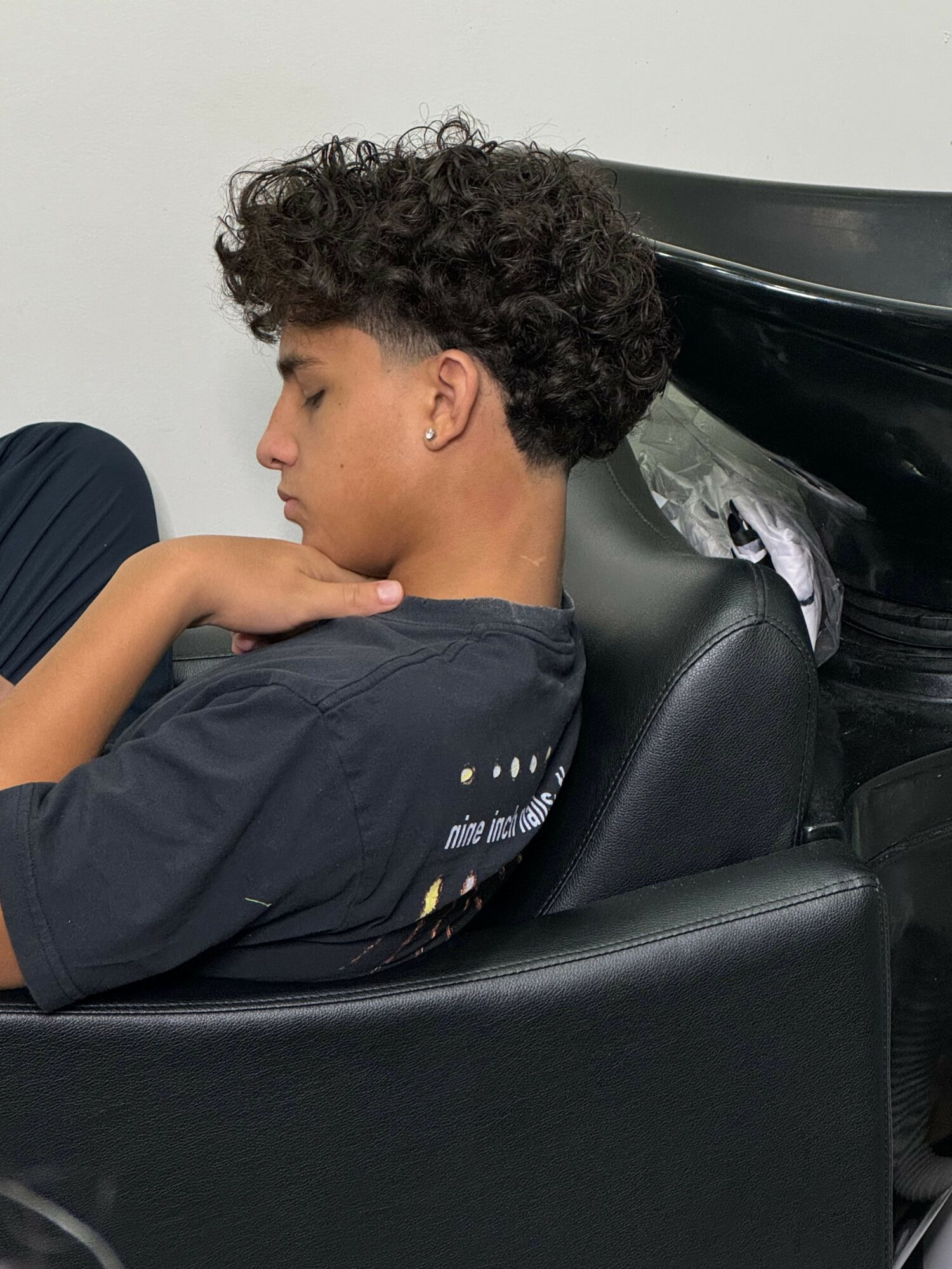 Young person with curly hair sitting in a car seat, head tilted downward, hand on neck, wearing a dark shirt.