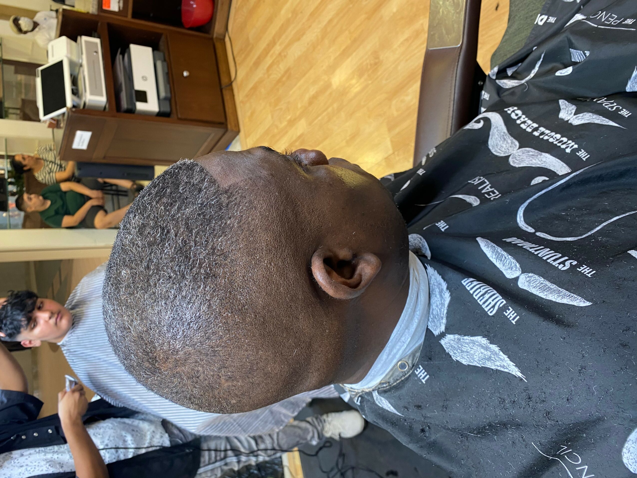 Person with a shaved head sitting in a barber chair, facing downward, in a barbershop with wooden floor and shelves.