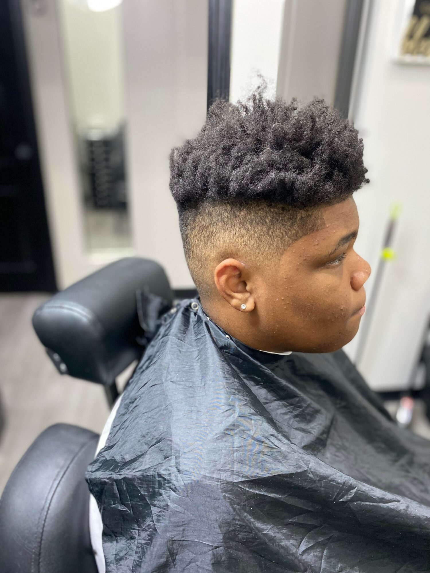 Young man with a textured, curly hairstyle, sitting in a barber chair, wearing a black cape, in a barbershop.