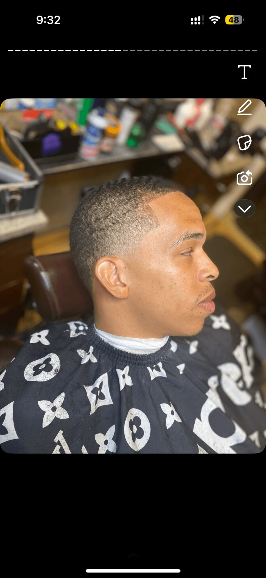Side profile of a man with short hair, sitting in a barber shop chair, wearing a patterned cape.