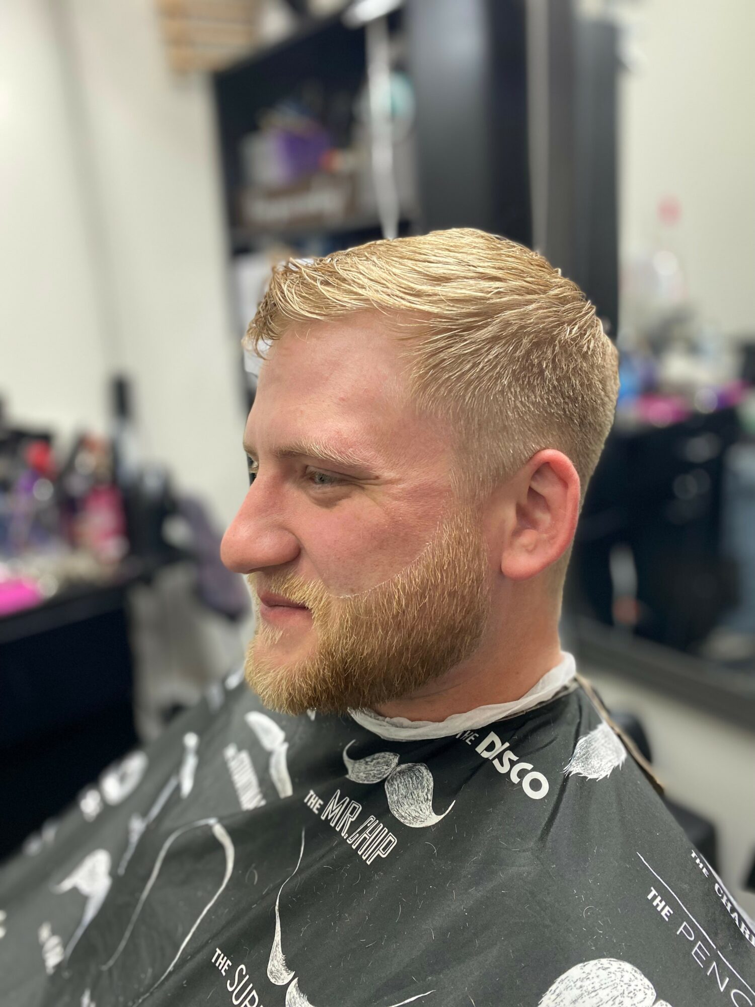 Side profile of a man with short blonde hair and a beard, sitting in a barbershop chair, smiling.