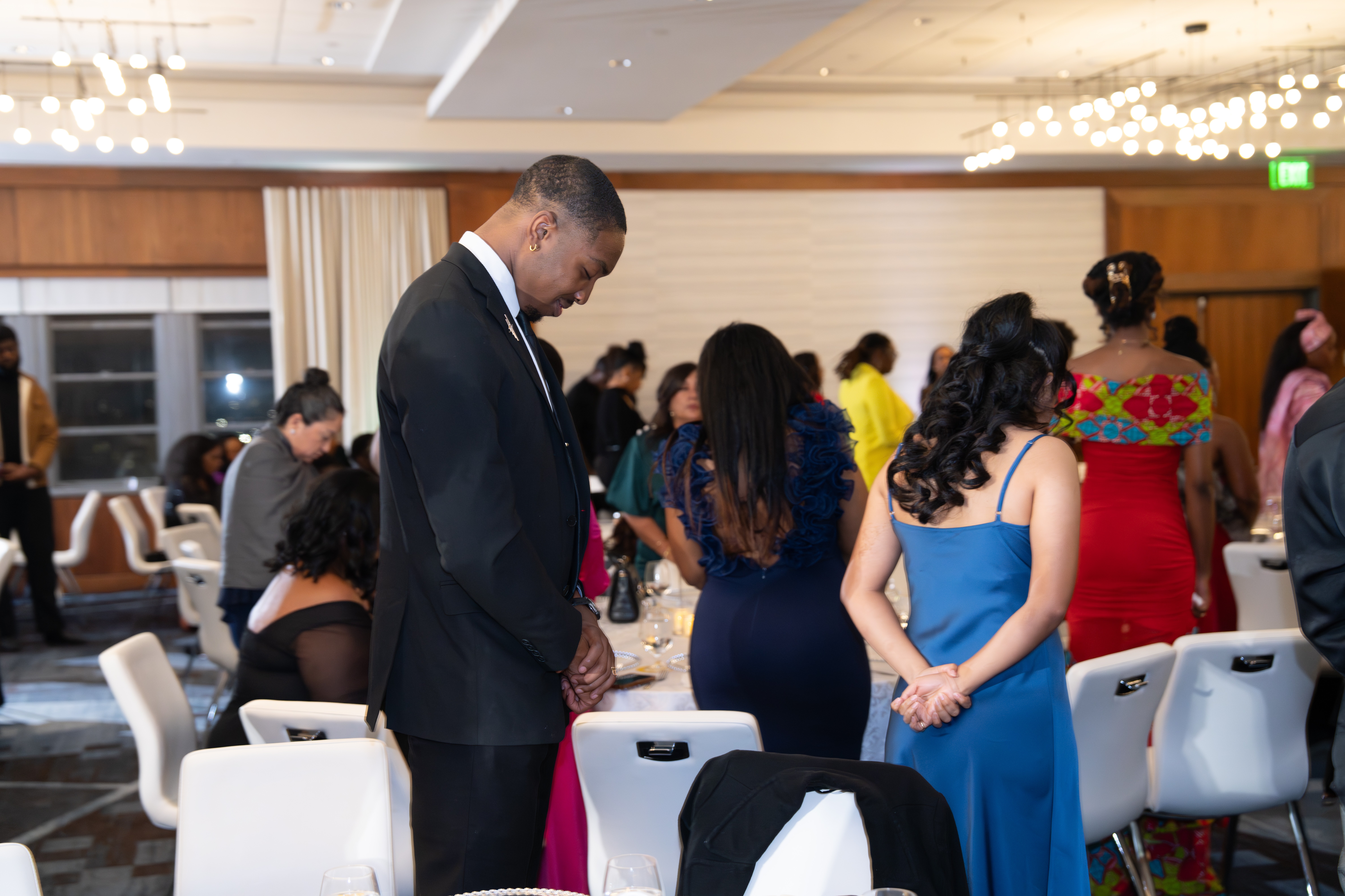 People gathered in a well-lit banquet hall, some standing and some seated, dressed in formal attire, engaging in conversation.