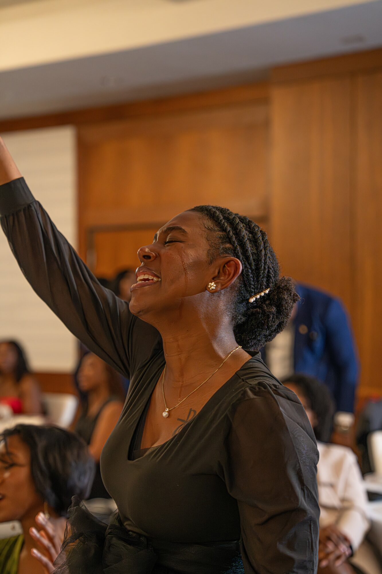Woman with braided hair smiling and raising her arm in a room with people and wooden paneling.