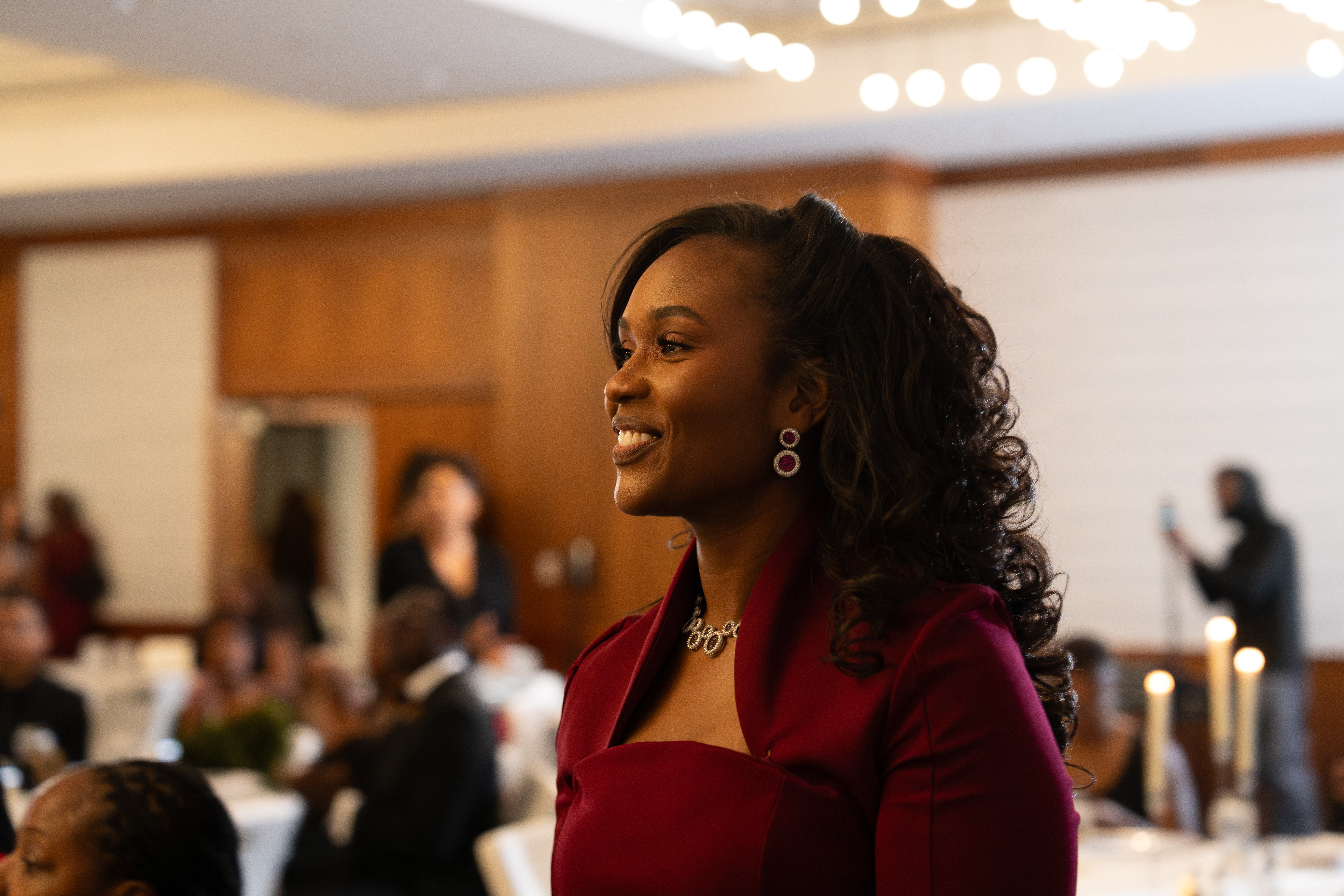 Smiling woman with curly hair wearing a red dress and jewelry at an indoor event, with people and decorations in background.