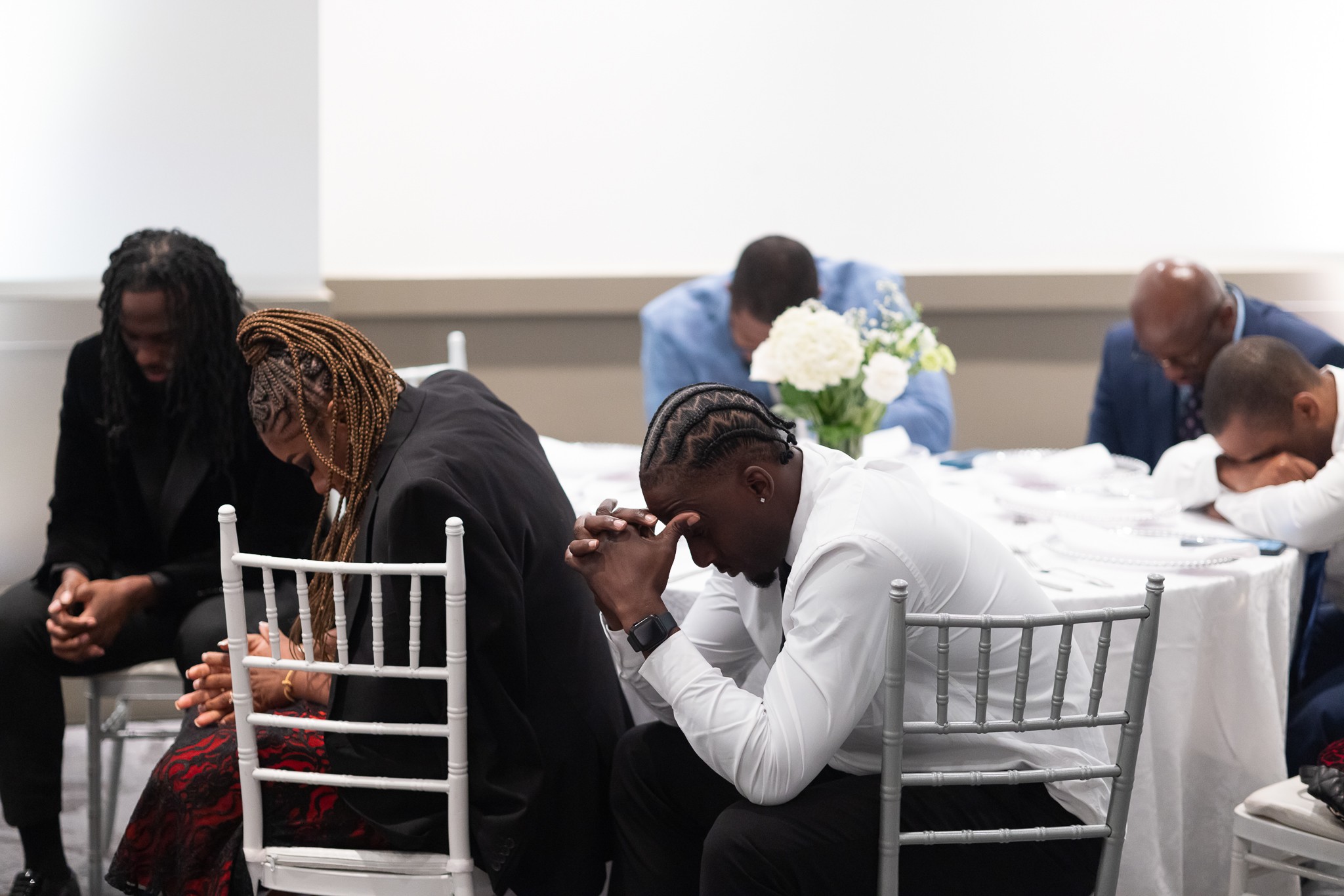 People sitting with heads bowed, hands clasped, in a prayer or reflection, in a bright room with white walls and flowers.