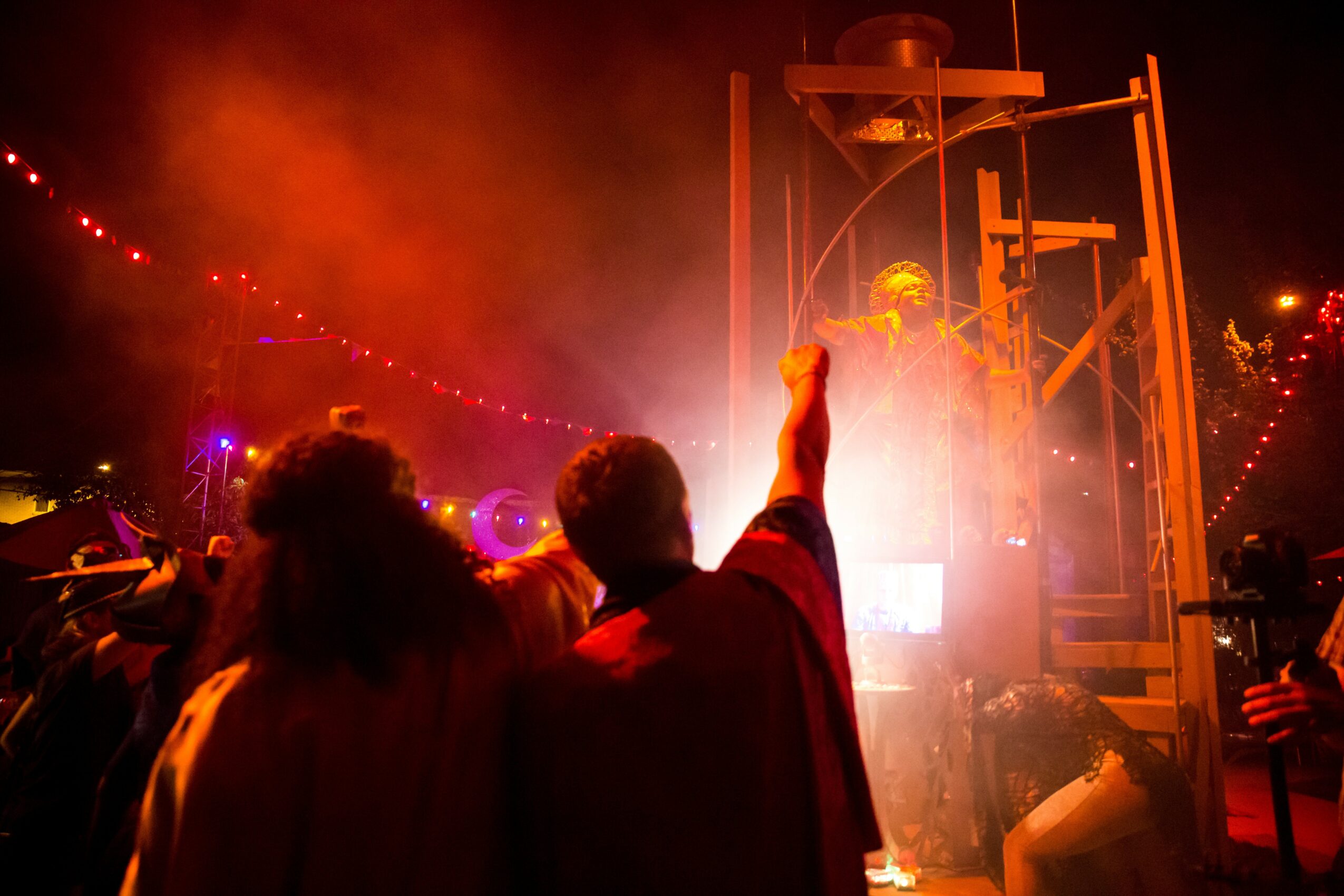 People at an outdoor event with colorful lights and a performer on a stage, some raising their hands, night scene.